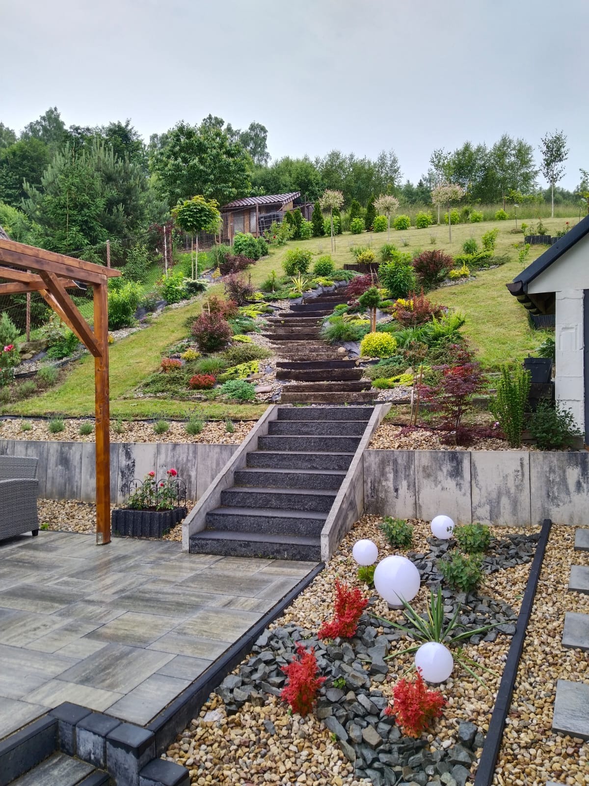Ukształtowany ogród na zboczu z kamiennymi schodami, rabatami kwiatowymi, ozdobnymi krzewami i kulistymi lampami ogrodowymi, widok z tarasu.