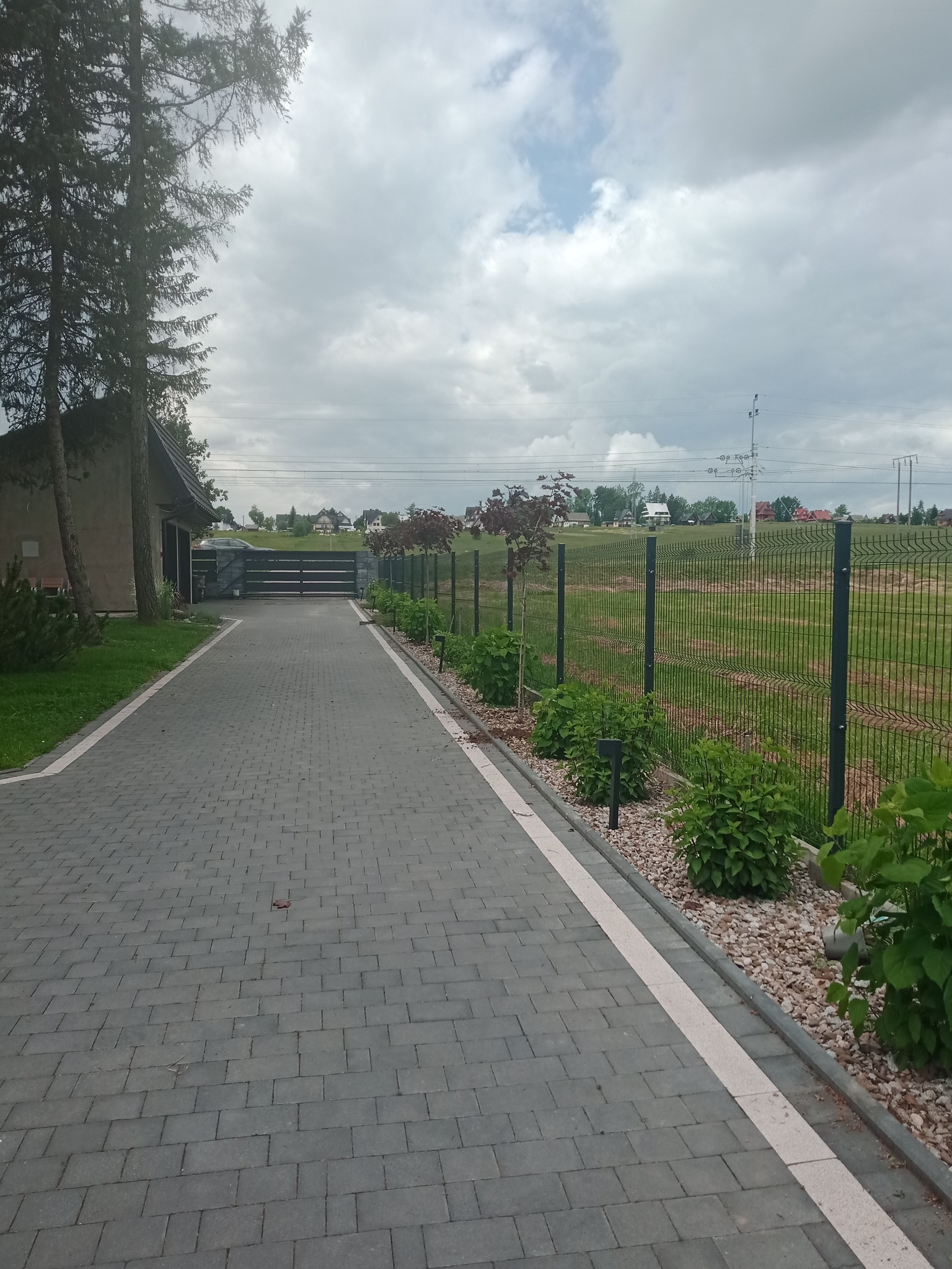 Wyłożona kostką brukową, szara alejka prowadzi do bramy wjazdowej, wzdłuż ogrodzenia z siatki posadzone krzewy i młode drzewka z bordowymi liśćmi, podświetlone niskimi lampami.