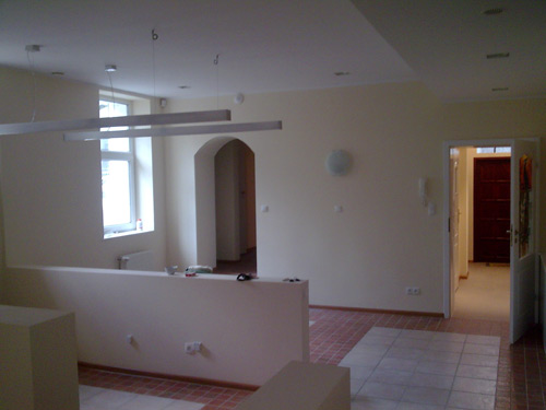 Wnętrze biura po remoncie: pomalowane ściany w odcieniach beżu, widoczne elementy instalacji elektrycznej, fragment podłogi z terakoty oraz drzwi do kolejnego pomieszczenia z drewnianą ościeżnicą.