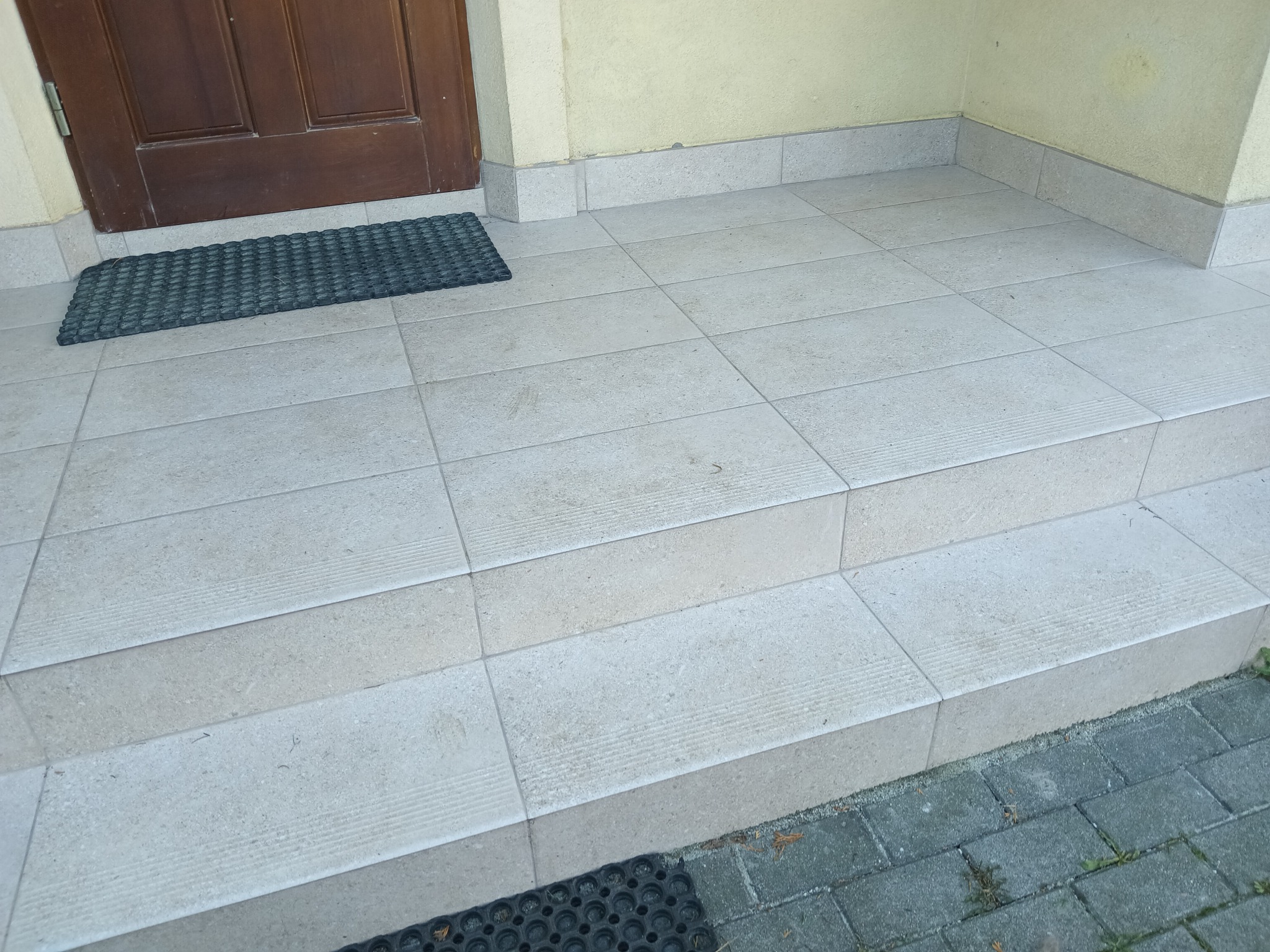 Wykończone schody wejściowe do domu z jasnych, kwadratowych płytek ceramicznych, z antypoślizgowymi rowkami na krawędziach stopni i gumową wycieraczką przed drzwiami.