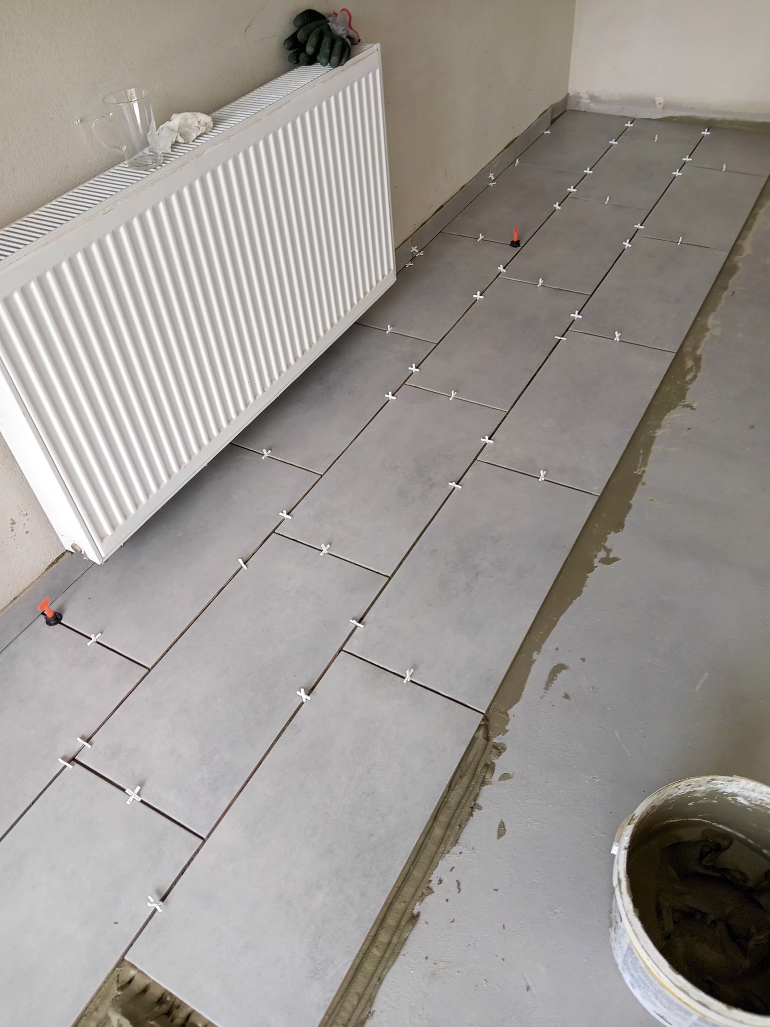 Układanie szarych płytek podłogowych z użyciem krzyżyków dystansowych, widoczny grzejnik, wiadro z zaprawą i rękawice robocze.