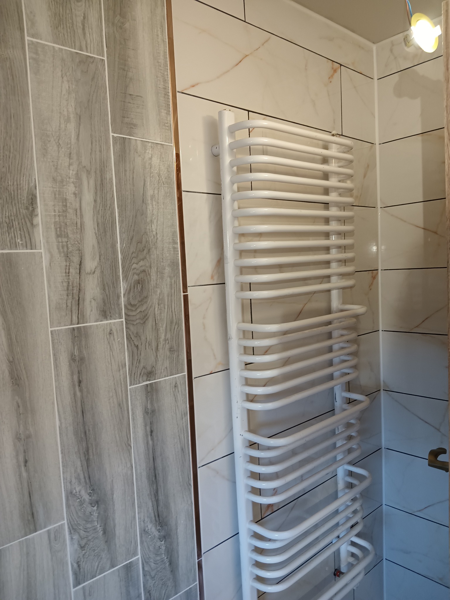Biały, drabinkowy grzejnik łazienkowy na tle płytek imitujących marmur i drewna w odcieniach bieli i szarości; widoczny fragment instalacji oświetleniowej w górnym rogu.