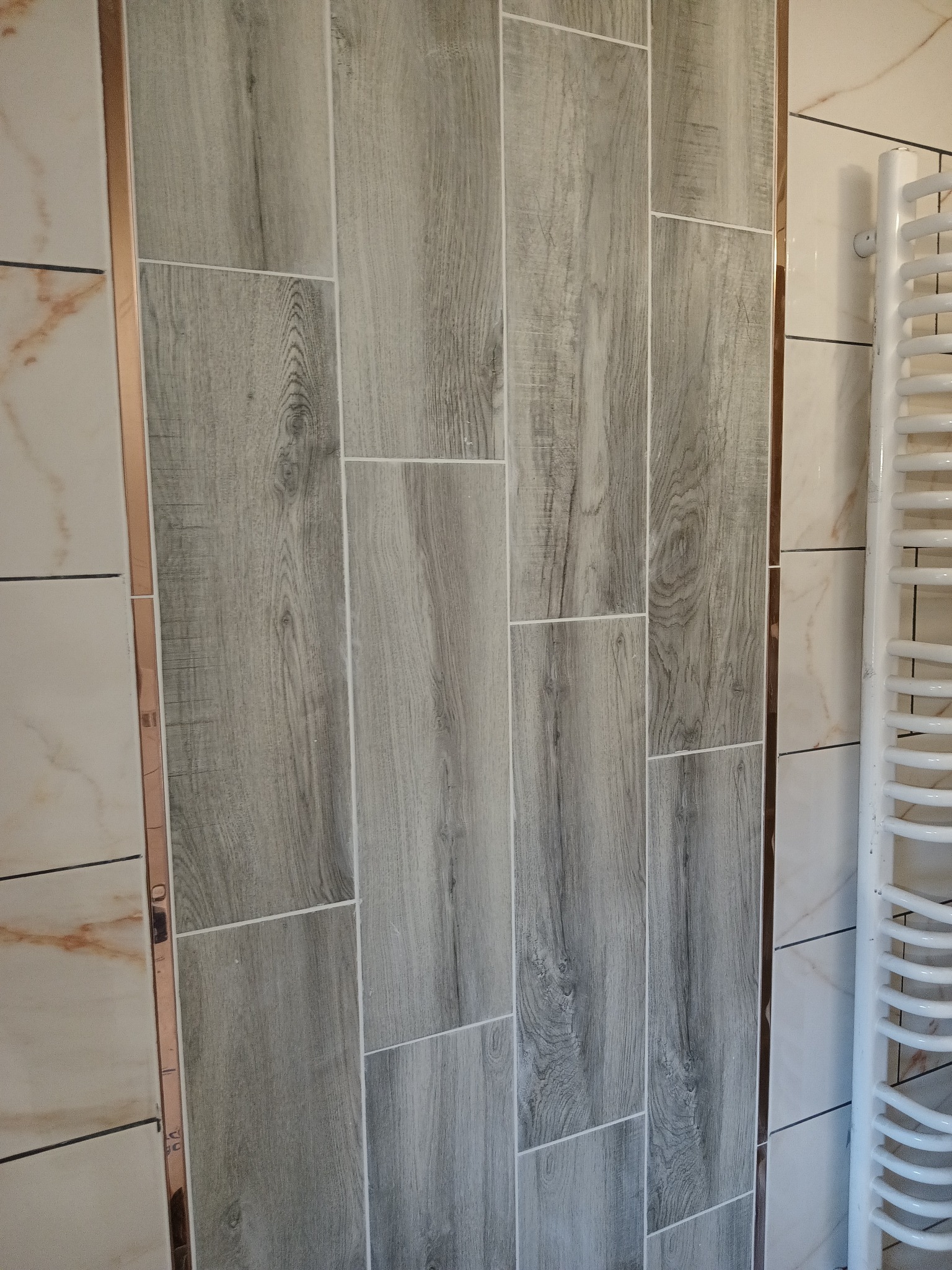 Ściana łazienki wyłożona płytkami imitującymi drewno w odcieniach szarości, z widocznym grzejnikiem drabinkowym i dekoracyjną listwą w kolorze miedzi.