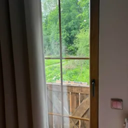 Drewniane okno z widokiem na zielony ogród, częściowo zasłonięte szarą zasłoną, z widocznym drewnianym balkonem i włącznikiem światła na ścianie.