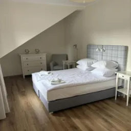 Jasna sypialnia na poddaszu z łóżkiem małżeńskim, białą komodą i drewnianą podłogą. Na łóżku ułożone białe ręczniki.