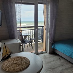 Efekt - Pokój z widokiem na morze, balkon z krzesłami i stolikiem, łóżko z błękitną narzutą, dekoracyjny model żaglówki na stole, pleciona podkładka. Widoczne ślady użytkowania, wymagające odświeżenia.