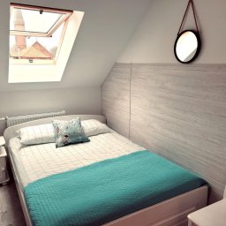 Efekt - Jasna sypialnia na poddaszu z oknem dachowym, widok na kościół, podwójne łóżko z turkusową narzutą i poduszką w morskim stylu, okrągłe lustro na ścianie z boazerią.