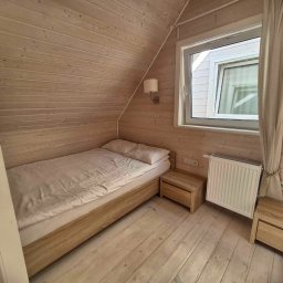 Efekt - Drewniany pokój z pojedynczym łóżkiem, stolikami nocnymi i grzejnikiem pod oknem, oświetlony lampą ścienną.