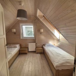 Efekt - Pokój na poddaszu w jasnej kolorystyce z dwoma pojedynczymi łóżkami, drewnianą podłogą i skośnym sufitem, widok zza uchylonych drzwi.