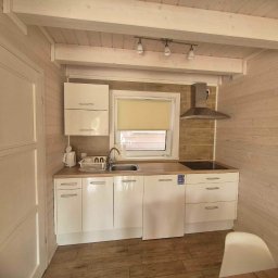 Efekt - Wnętrze jasnej kuchni w domku letniskowym z drewnianymi ścianami, białymi szafkami, zlewozmywakiem i płytą grzewczą, oświetlone reflektorami.