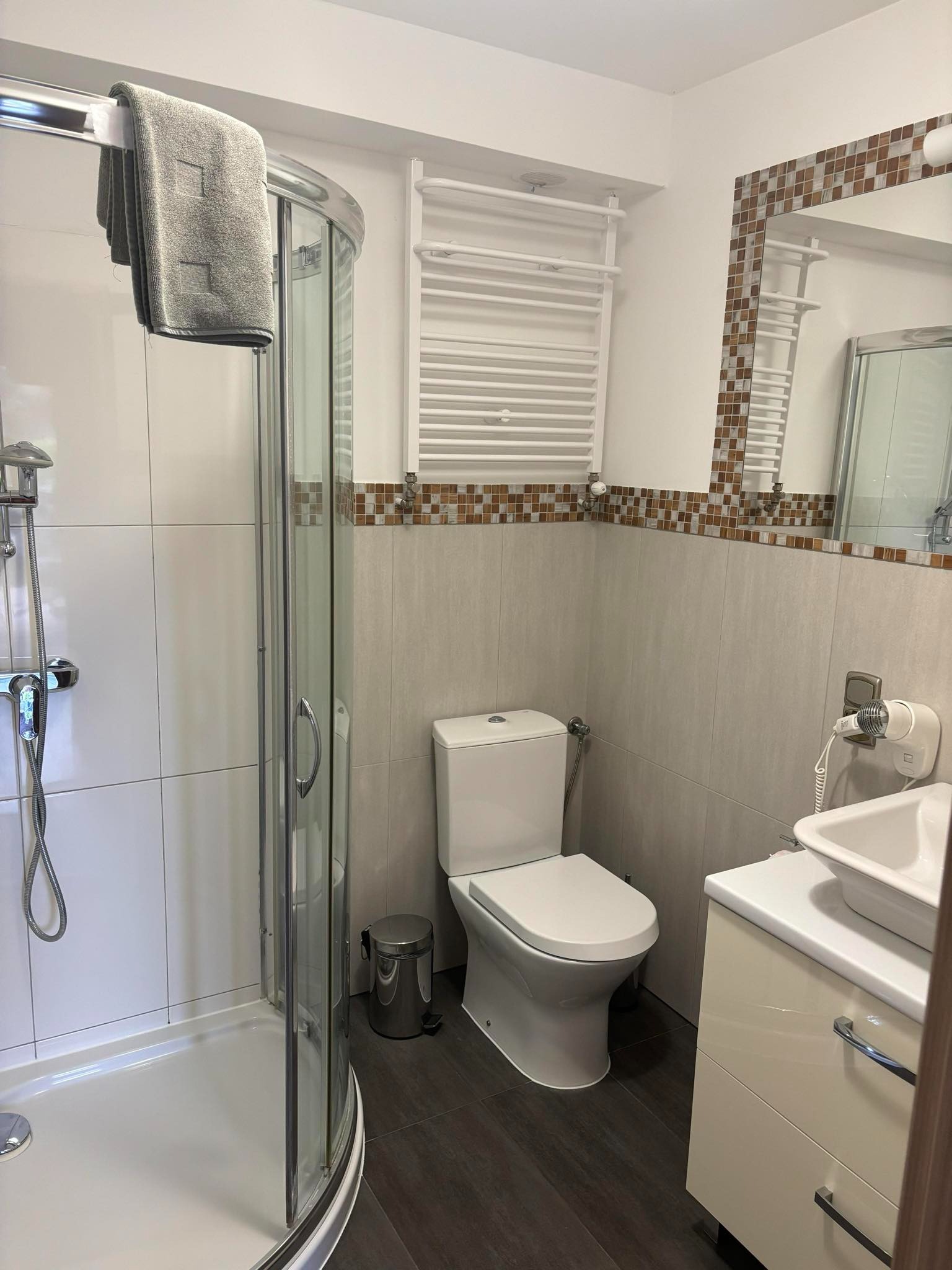 Jasna łazienka z kabiną prysznicową, toaletą, umywalką z szafką, grzejnikiem drabinkowym i lustrem z mozaikową ramą.