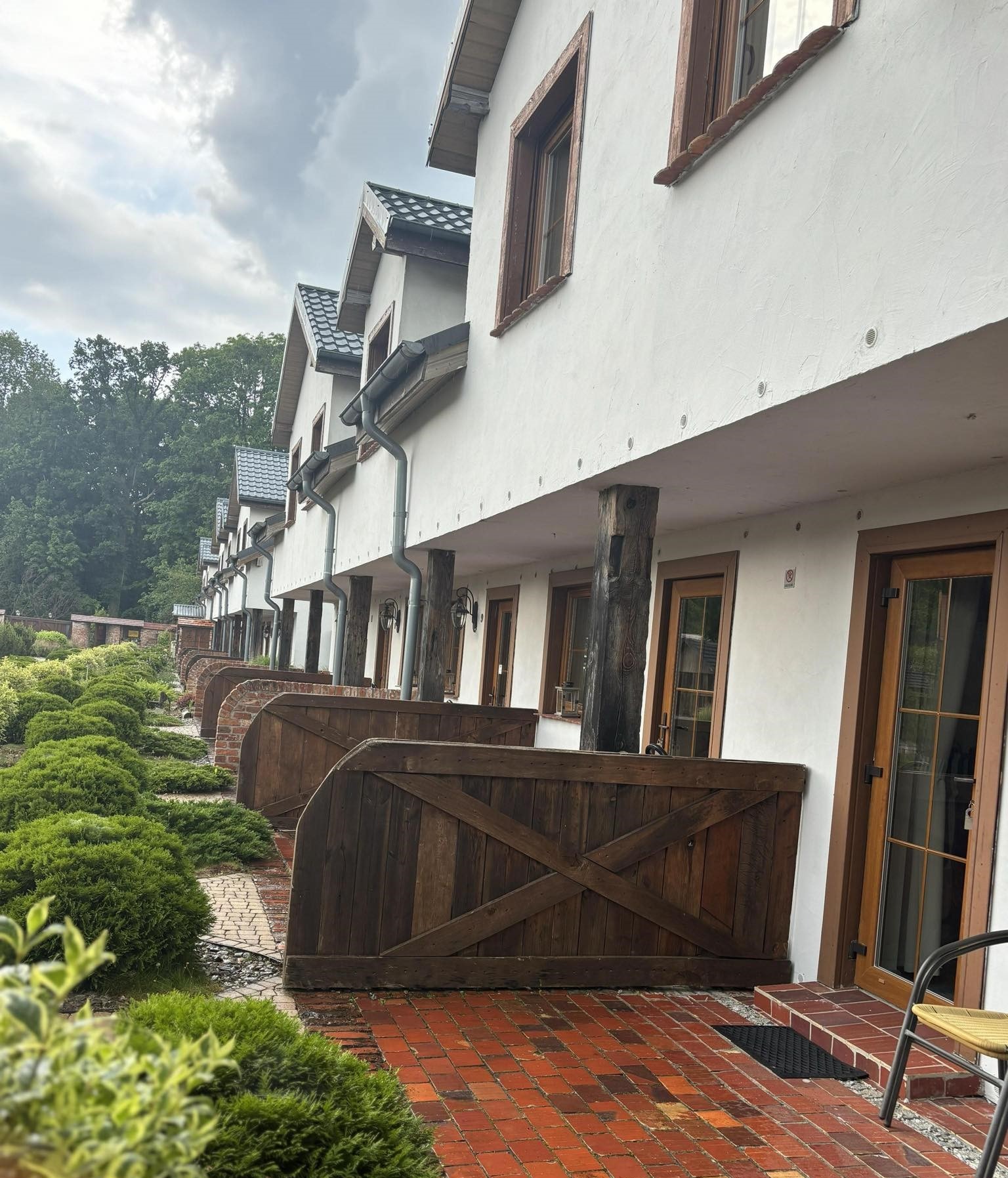 Rząd białych budynków z drewnianymi oknami, kolumnami i balustradami w stylu rustykalnym, widok z zewnątrz na zadbany ogród z niskimi krzewami i ceglanym chodnikiem.