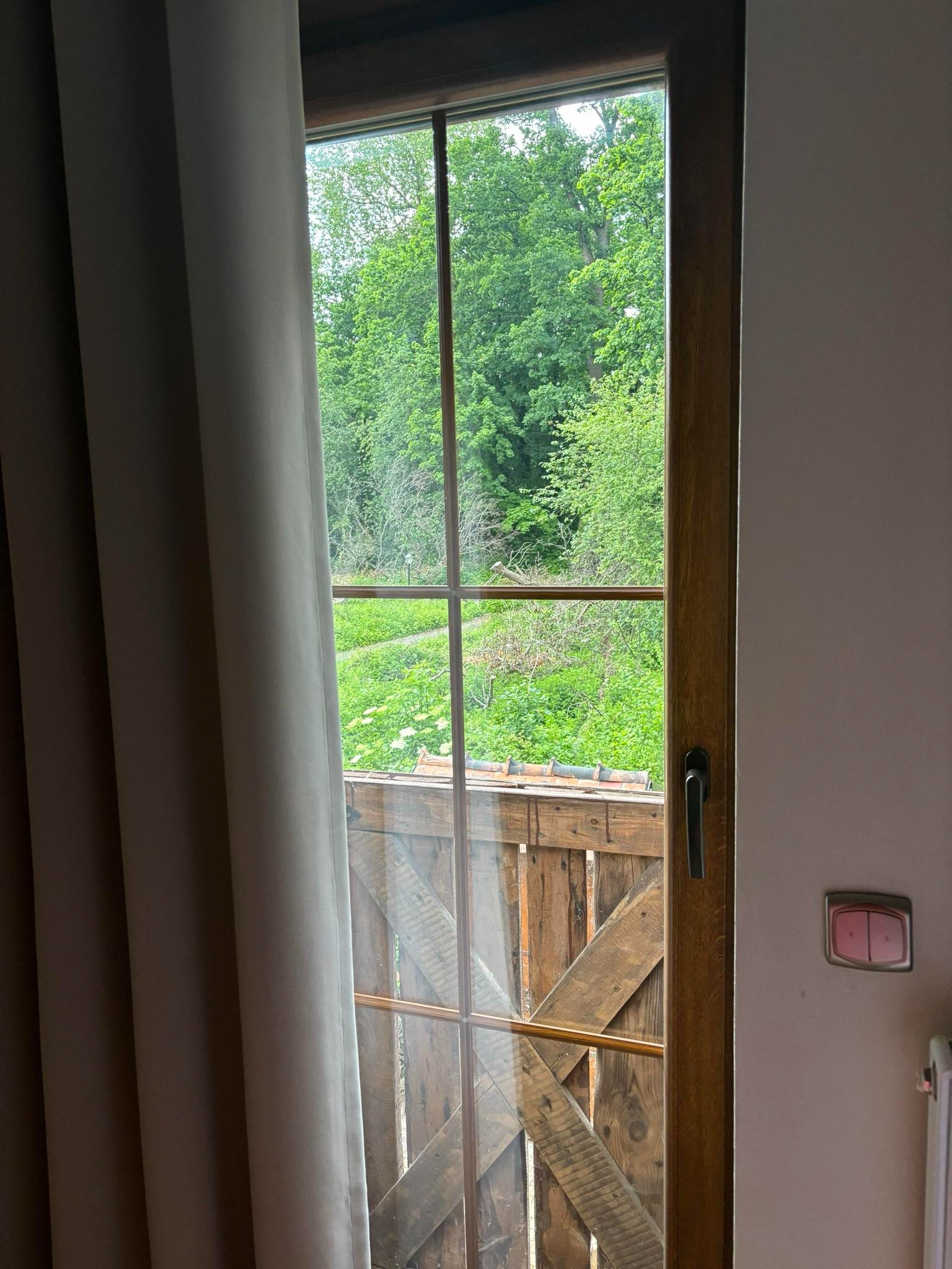 Drewniane okno z widokiem na zielony ogród, częściowo zasłonięte szarą zasłoną, z widocznym drewnianym balkonem i włącznikiem światła na ścianie.