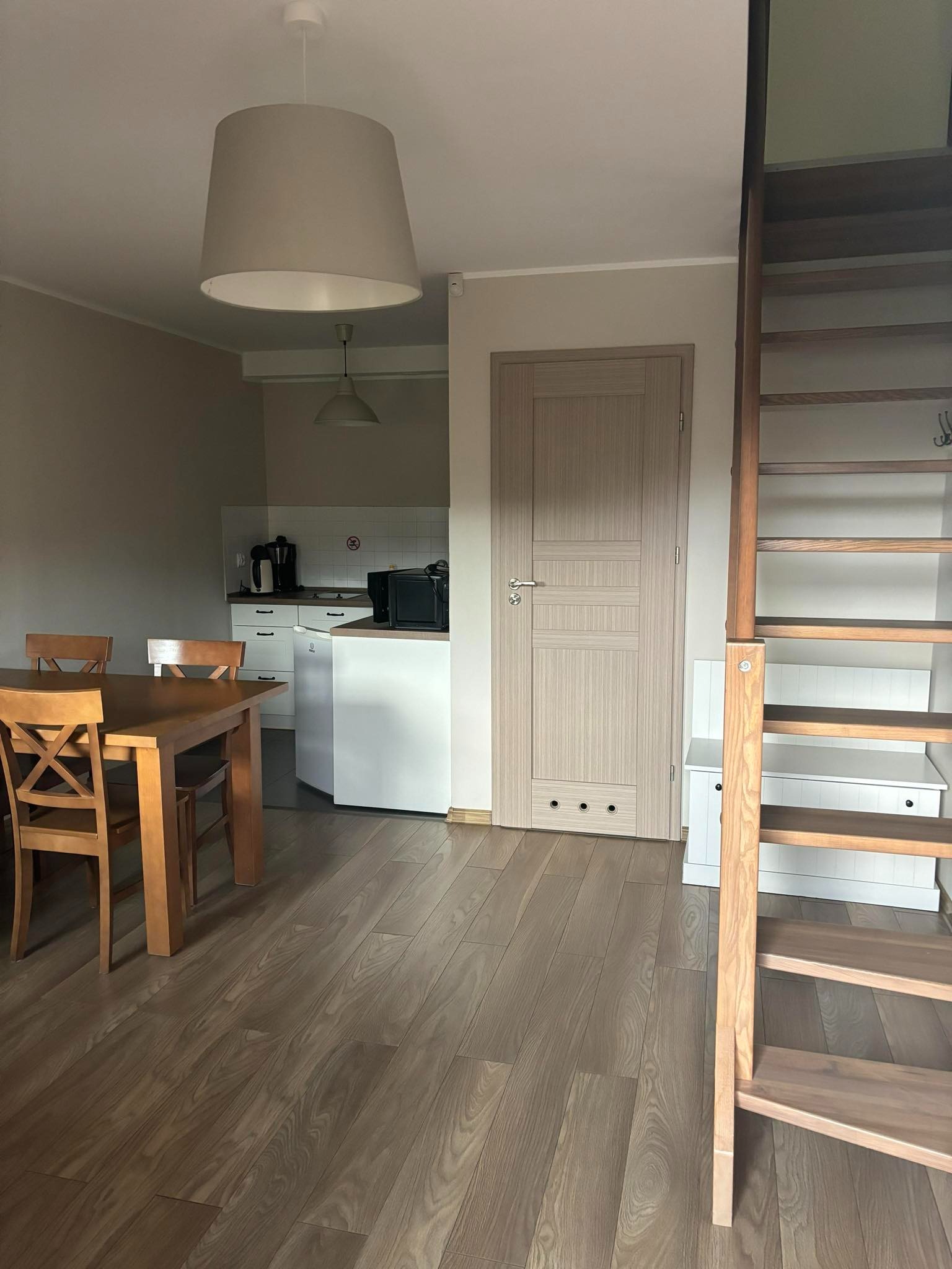 Jasne wnętrze mieszkania z drewnianym stołem, krzesłami, schodami drabinkowymi i aneksem kuchennym z białymi szafkami.