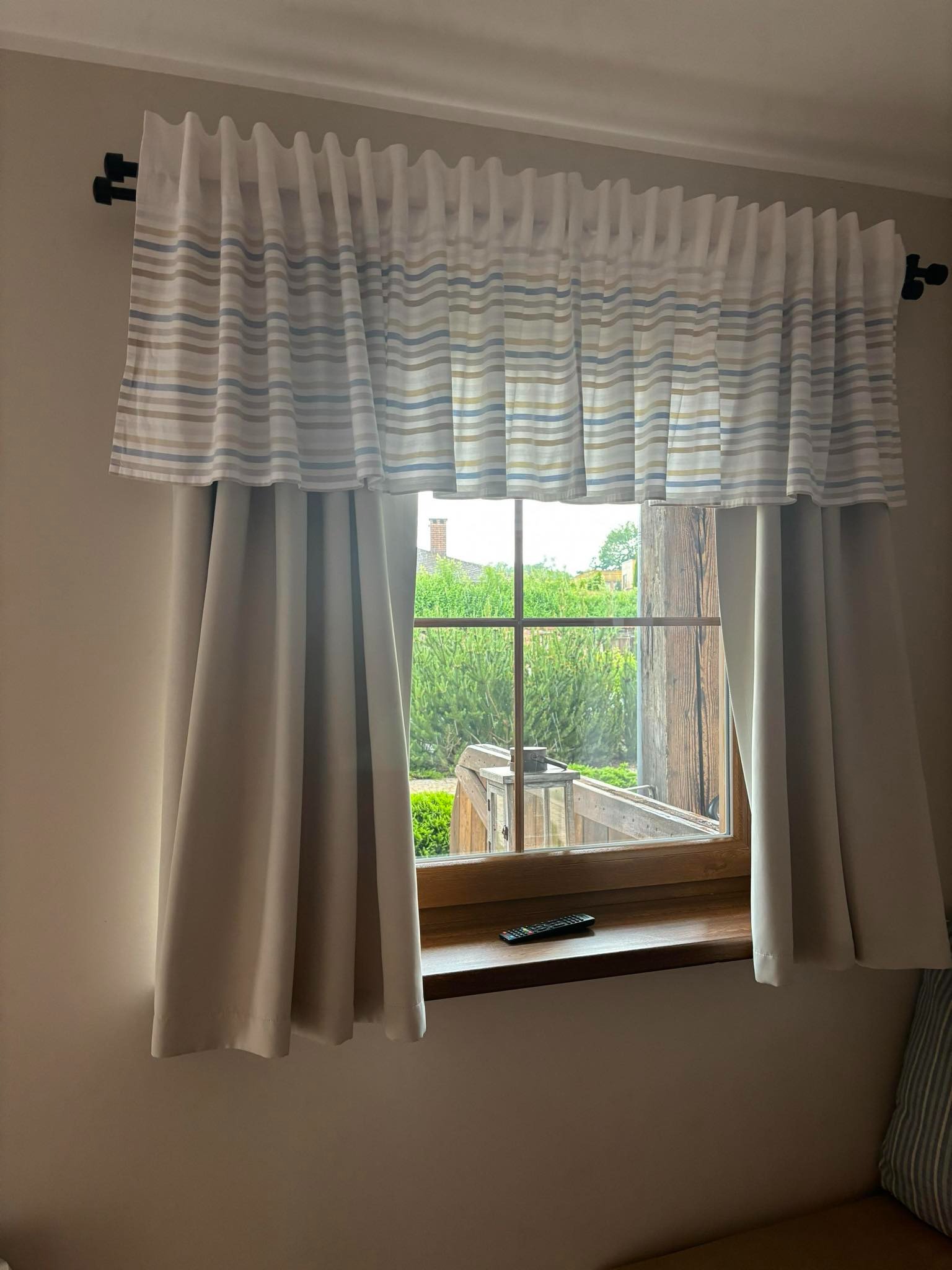 Okno z drewnianą ramą i zasłonami w paski, widok na zielony ogród z zewnątrz, pilot na parapecie.