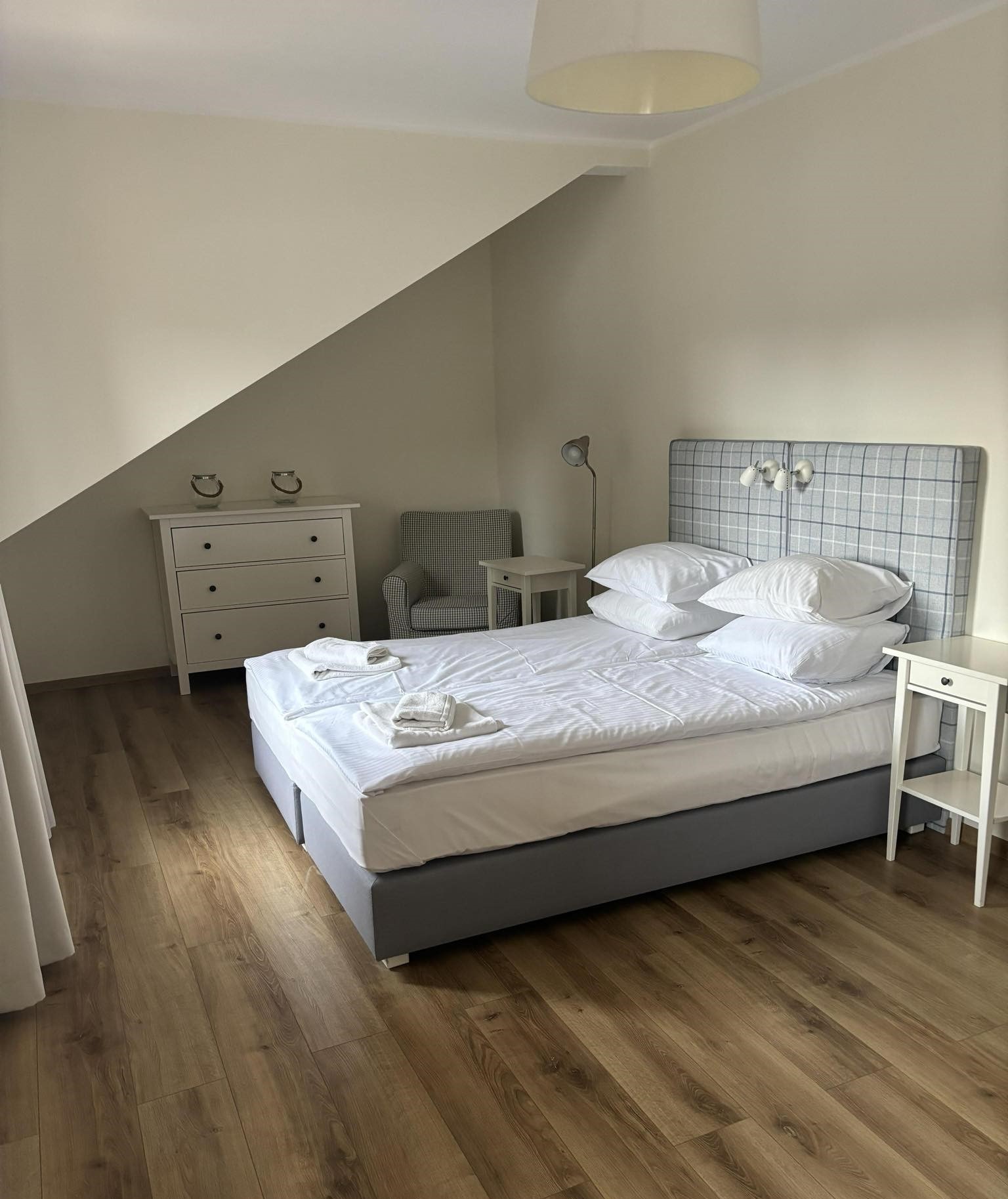 Jasna sypialnia na poddaszu z łóżkiem małżeńskim, białą komodą i drewnianą podłogą. Na łóżku ułożone białe ręczniki.