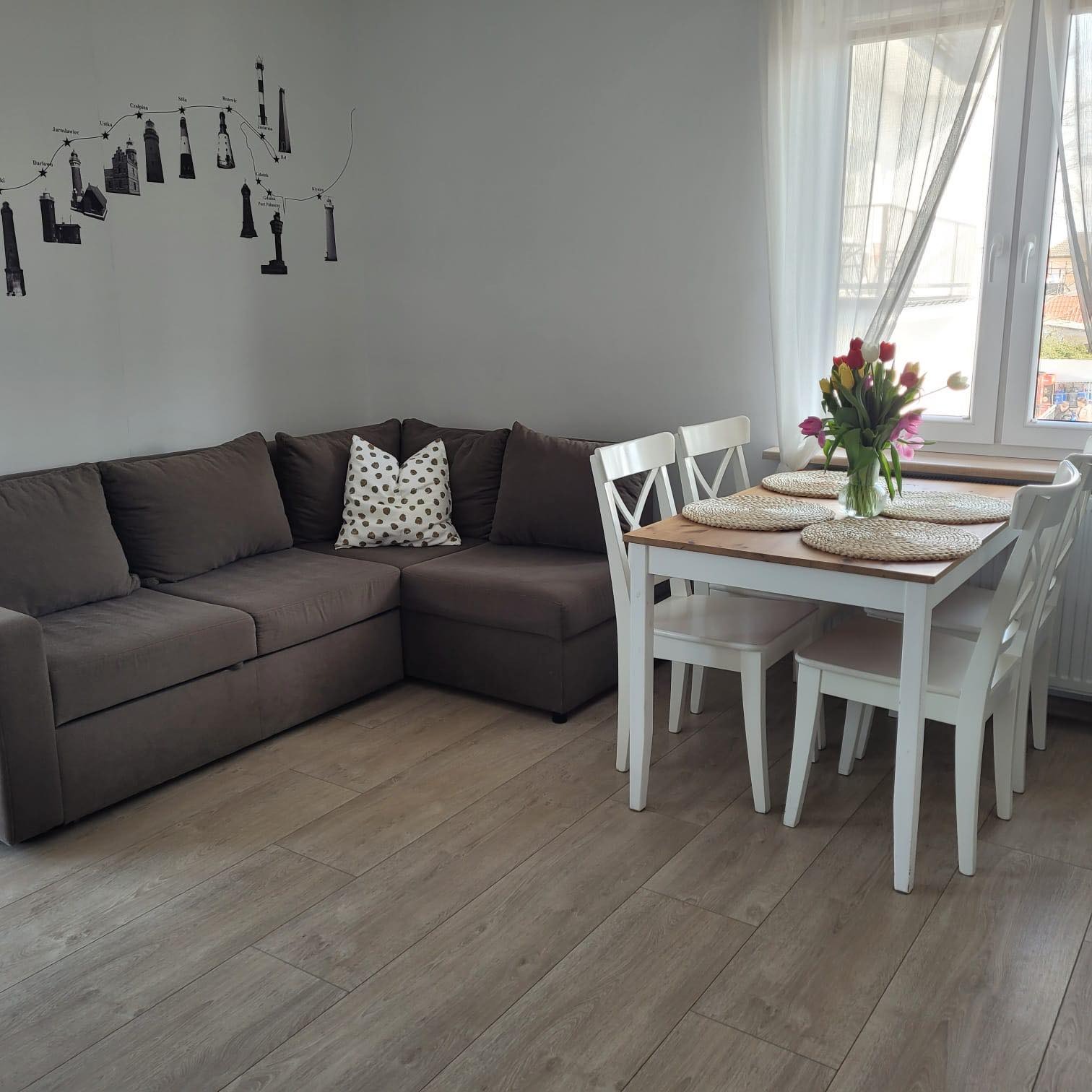 Salon z narożnikiem, białym stołem z bukietem tulipanów i dekoracją ścienną przedstawiającą latarnie morskie.