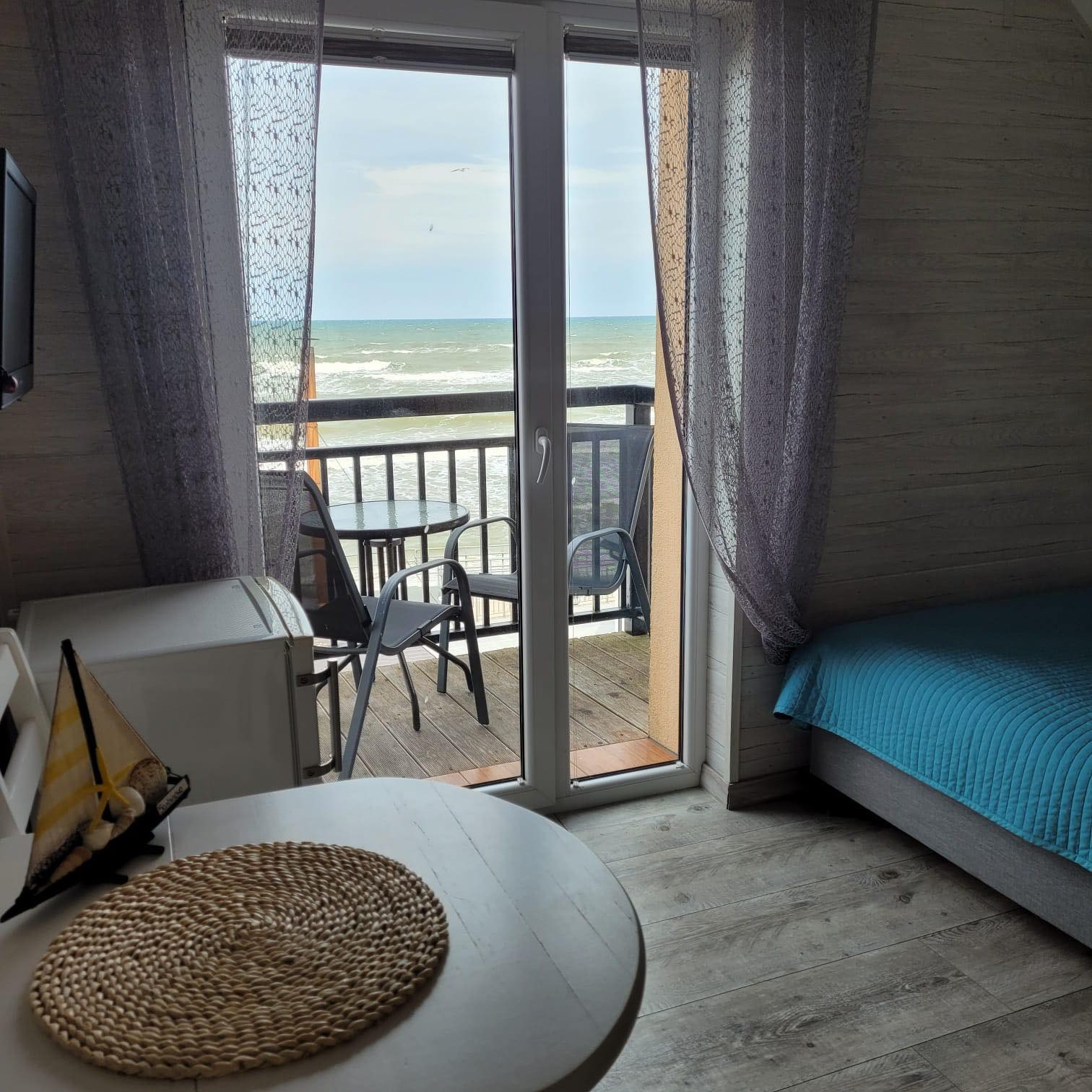 Pokój z widokiem na morze, balkon z krzesłami i stolikiem, łóżko z błękitną narzutą, dekoracyjny model żaglówki na stole, pleciona podkładka. Widoczne ślady użytkowania, wymagające odświeżenia.