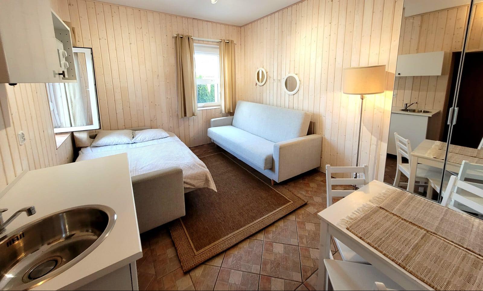 Jasne wnętrze pokoju z drewnianą boazerią, rozkładaną sofą, łóżkiem, stołem z krzesłami i aneksem kuchennym z umywalką.