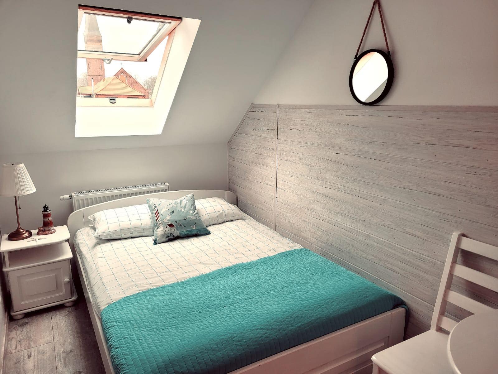 Jasna sypialnia na poddaszu z oknem dachowym, widok na kościół, podwójne łóżko z turkusową narzutą i poduszką w morskim stylu, okrągłe lustro na ścianie z boazerią.