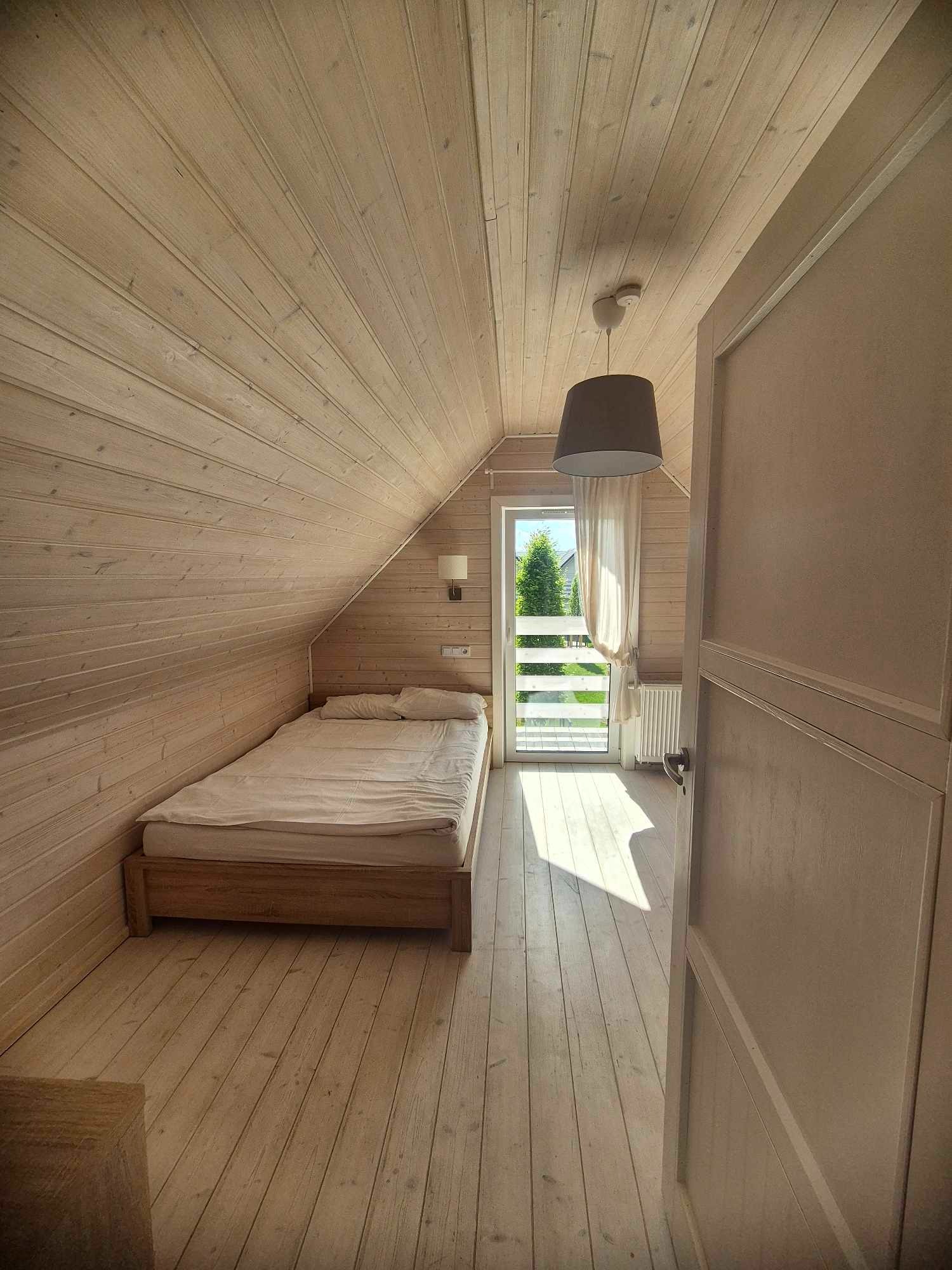 Jasne wnętrze pokoju na poddaszu z drewnianą boazerią, łóżkiem i wyjściem na balkon, widok przez uchylone drzwi.