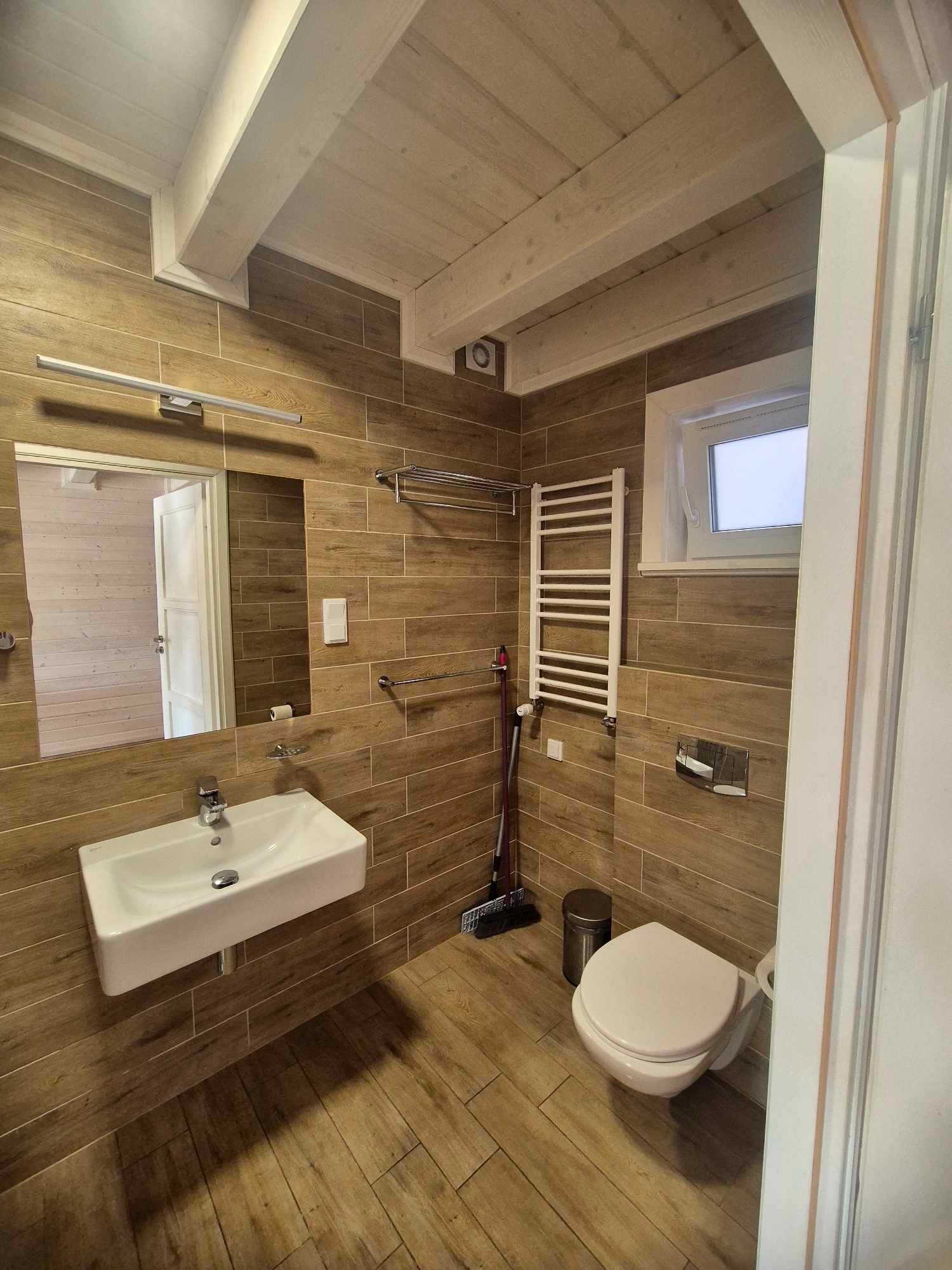 Jasna łazienka w brązowych płytkach imitujących drewno, z białą umywalką, toaletą i grzejnikiem drabinkowym, widoczna miotła i kosz na śmieci.