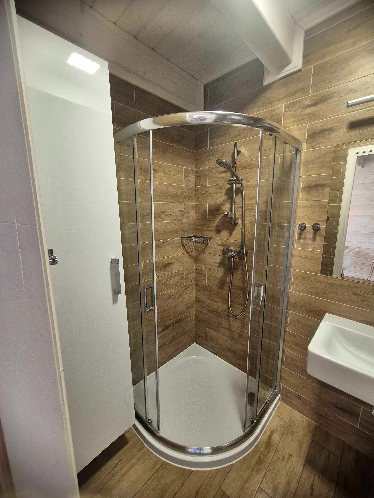 Nowoczesna kabina prysznicowa z chromowanymi profilami i deszczownicą na tle drewnopodobnych płytek, z widoczną umywalką i lustrem.