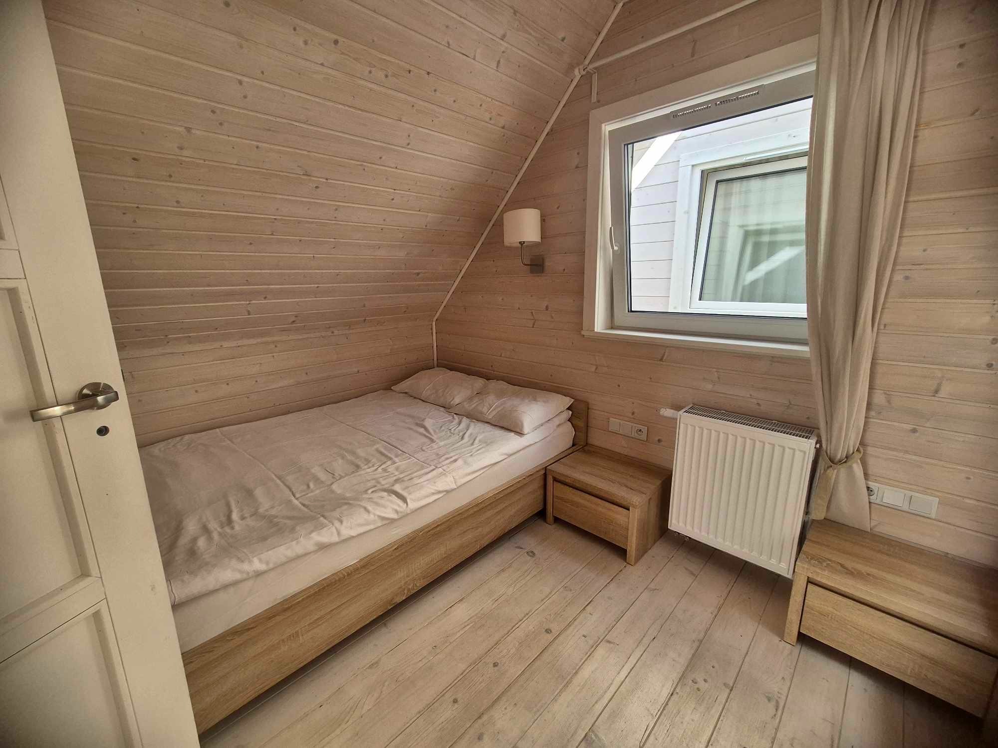 Drewniany pokój z pojedynczym łóżkiem, stolikami nocnymi i grzejnikiem pod oknem, oświetlony lampą ścienną.