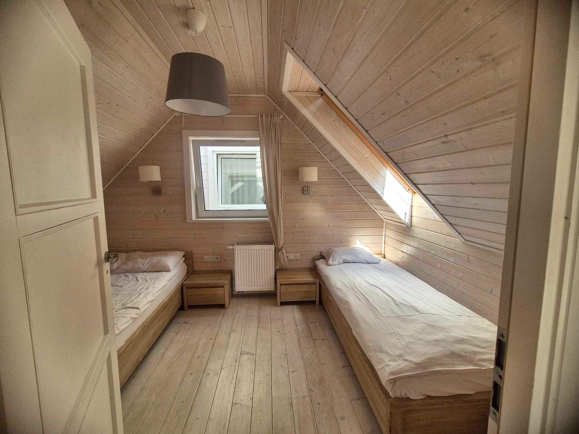 Pokój na poddaszu w jasnej kolorystyce z dwoma pojedynczymi łóżkami, drewnianą podłogą i skośnym sufitem, widok zza uchylonych drzwi.