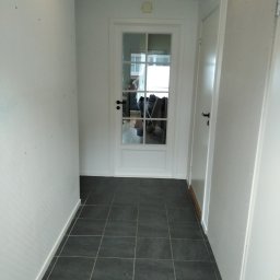 PMW - Korytarz z ciemnymi płytkami podłogowymi, białymi ścianami i sufitem z drewnianą boazerią, widok na białe drzwi z szybami i kolejne drzwi po prawej stronie.