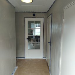 PMW - Korytarz w trakcie malowania, ściany zabezpieczone taśmą malarską, widoczna biała stolarka drzwiowa i sufit z panelami.