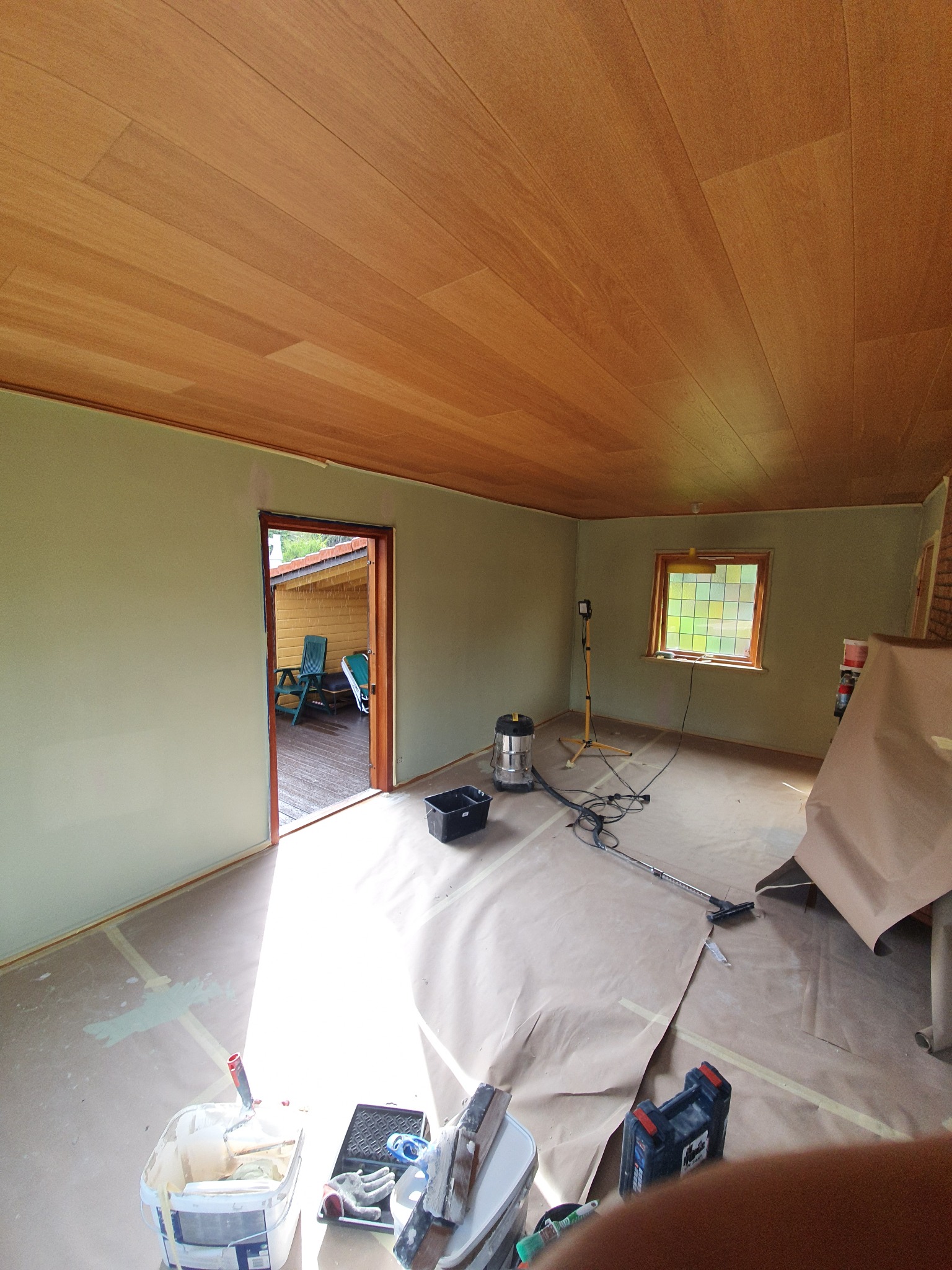 Pokój w trakcie malowania, zabezpieczona podłoga, drewniany sufit, widoczne narzędzia malarskie i lampa oświetleniowa.