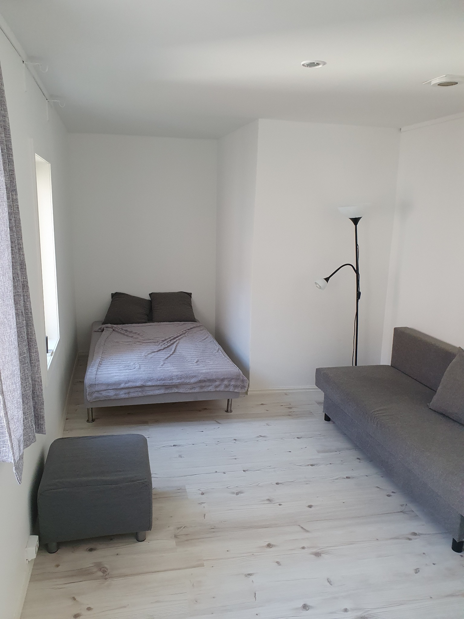 Jasne, minimalistyczne wnętrze pokoju z szarą sofą, łóżkiem z dwoma poduszkami, pufą i lampą podłogową, na podłodze widoczne jasne deski.