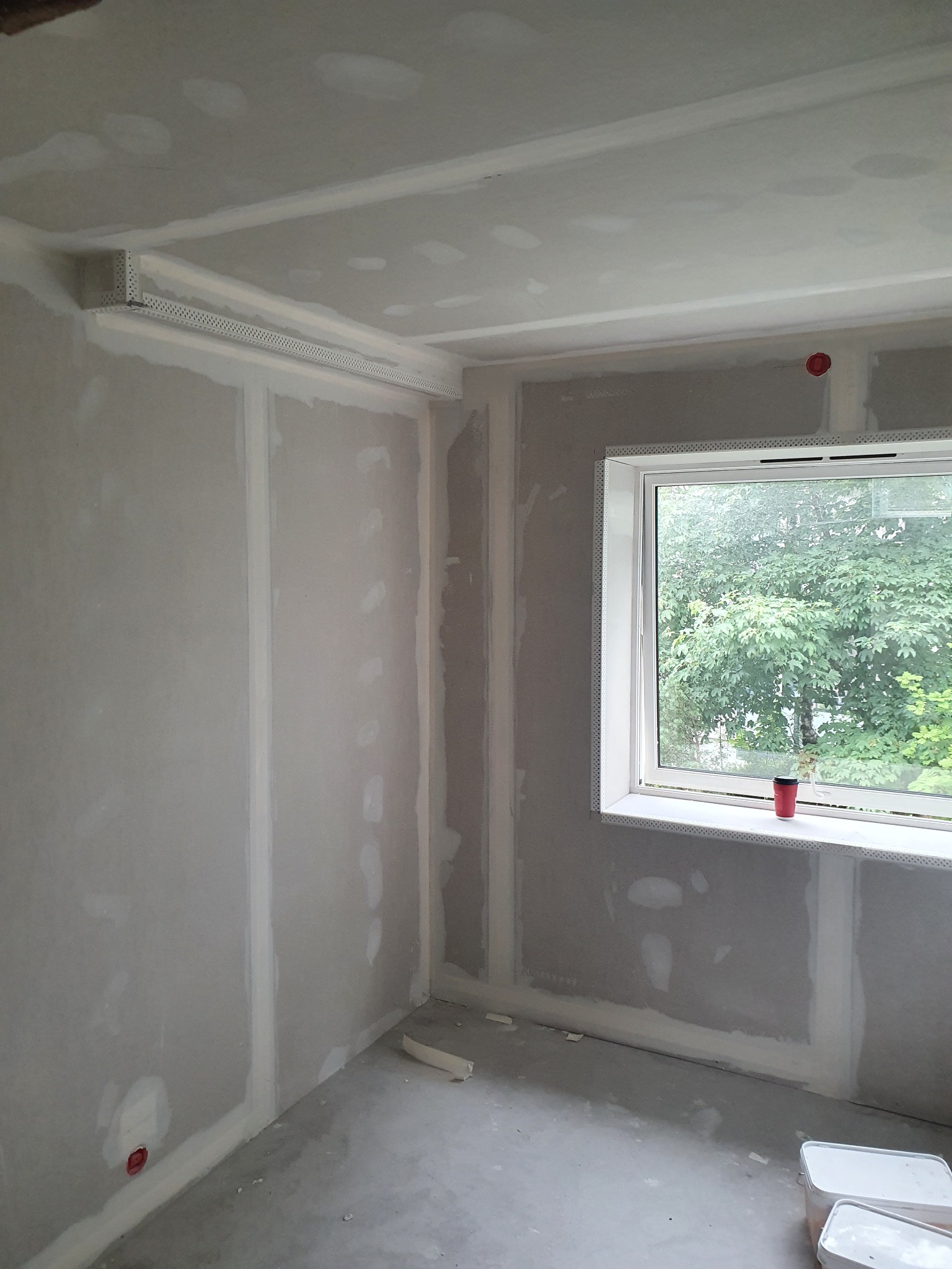 Pokój w trakcie prac wykończeniowych, widoczne ściany z płyt kartonowo-gipsowych, narożniki zabezpieczone profilami aluminiowymi, surowy betonowy sufit i podłoga, okno z widokiem na zieleń.