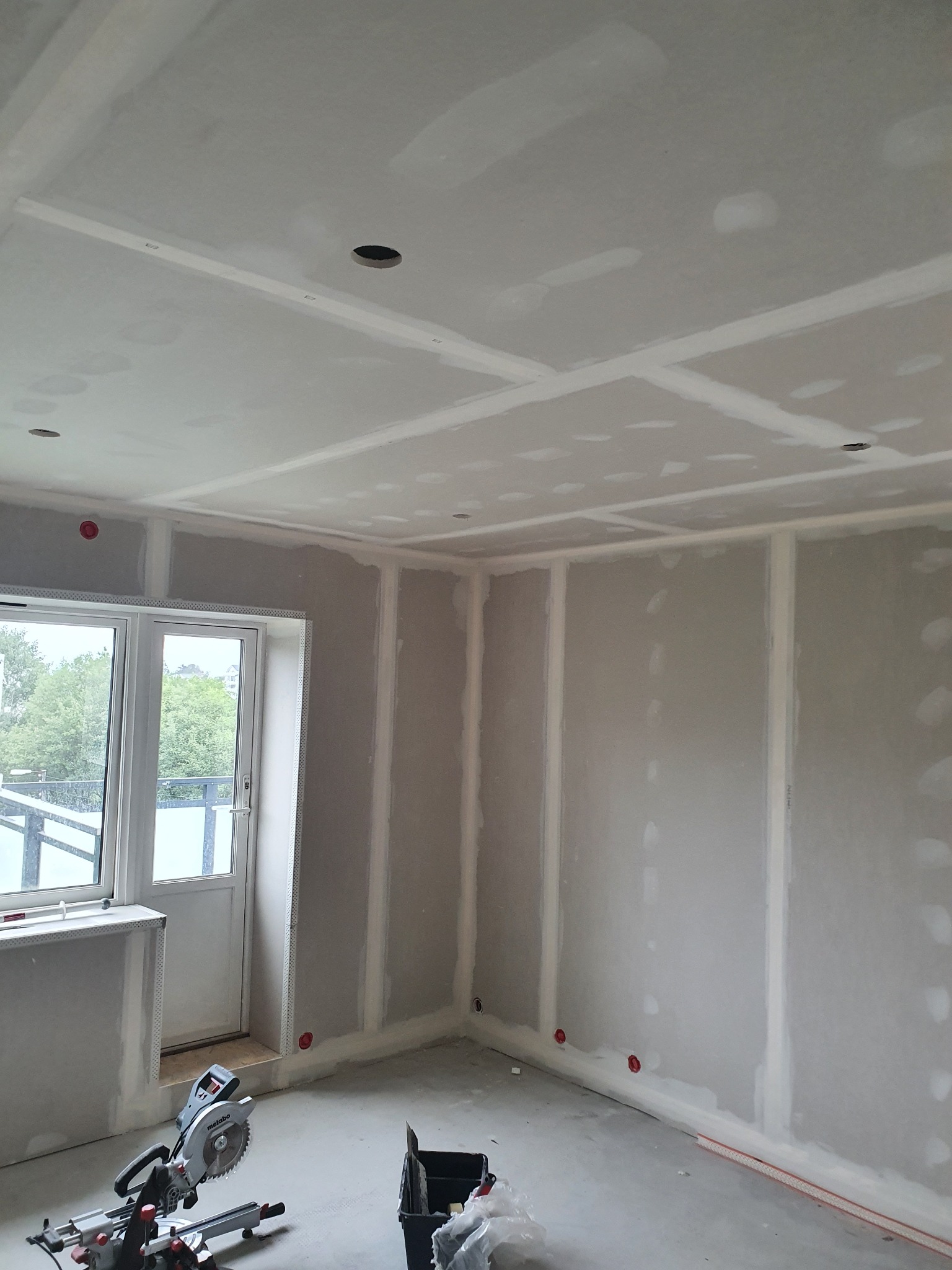 Pokój w trakcie montażu płyt kartonowo-gipsowych na ścianach i suficie, widoczne otwory na oświetlenie, szpachlowane łączenia, oraz piła ukośnica Metabo na podłodze.