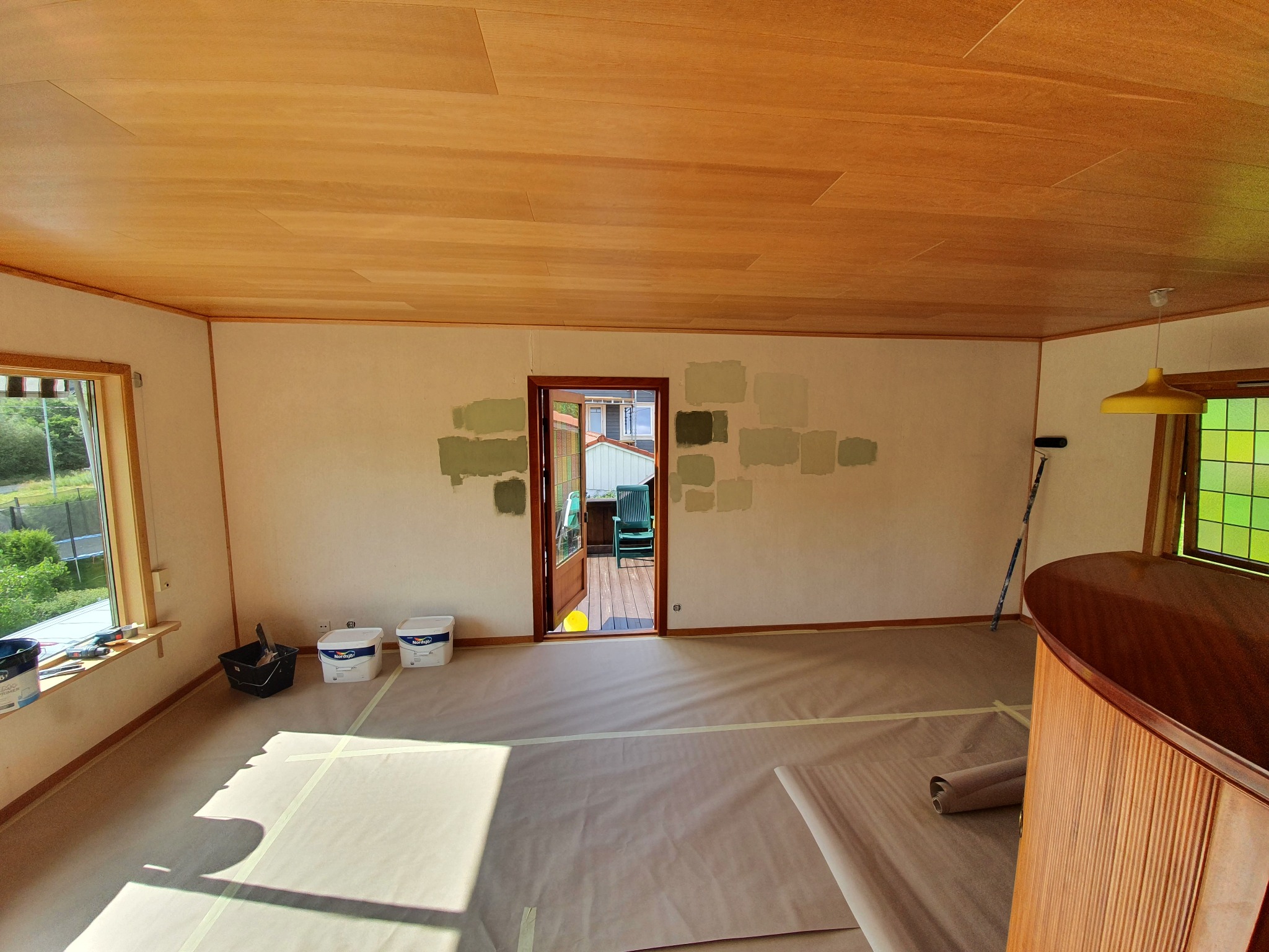 Pokój przygotowany do malowania: ściany z próbkami farb w odcieniach zieleni, podłoga i meble zabezpieczone folią malarską, widoczne pędzle, wałek i pojemniki z farbą.