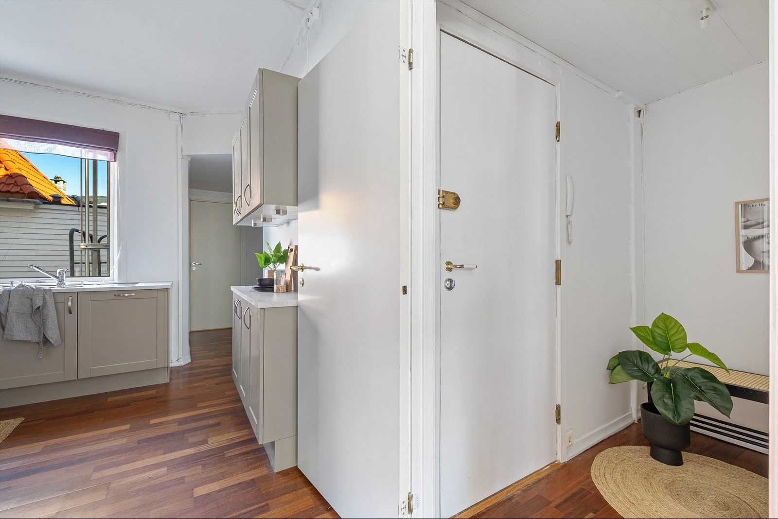 Wnętrze mieszkania z drewnianą podłogą, szarą zabudową kuchenną, białymi ścianami i drzwiami, widok na dach budynku za oknem.