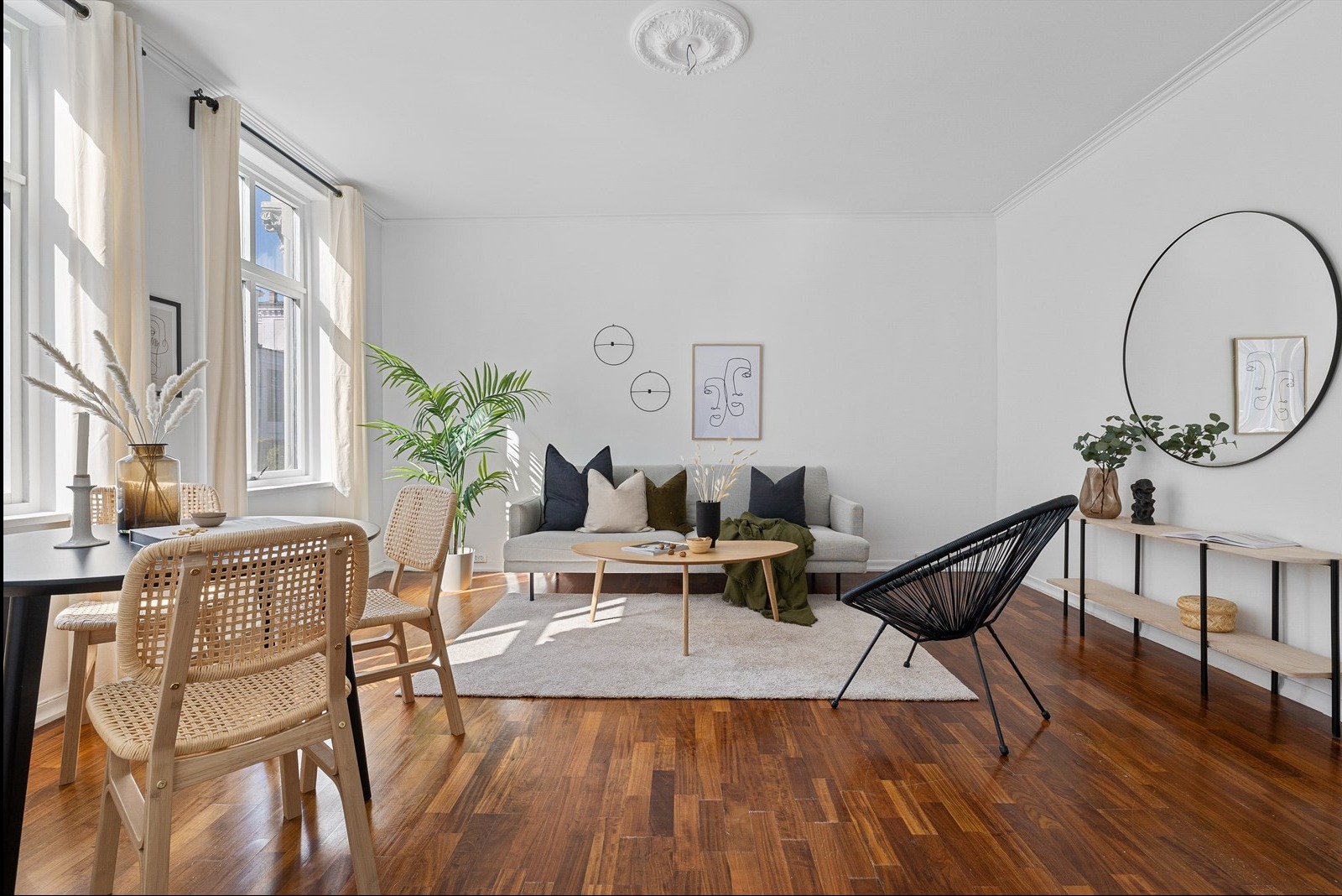 Jasne wnętrze salonu z drewnianą podłogą, beżowym dywanem, szarą sofą z poduszkami, okrągłym stolikiem kawowym, rattanowymi krzesłami, czarnym krzesłem z plecionki, roślinami w doniczkach...