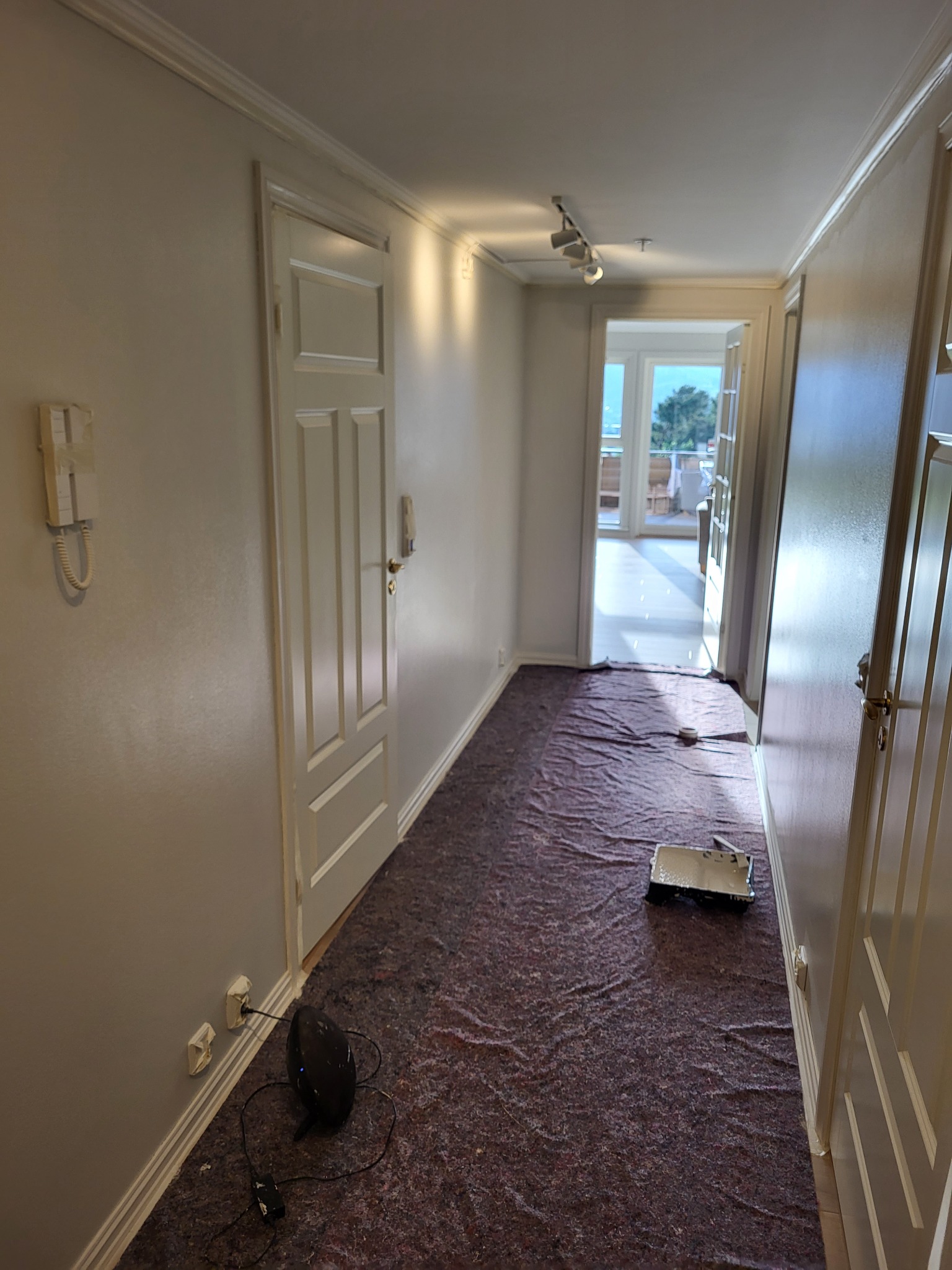 Korytarz w trakcie odświeżania, zabezpieczony podłogową folią ochronną, widoczna kuweta malarska z resztkami farby, ściany świeżo pomalowane na jasny odcień, w tle widok na balkon.