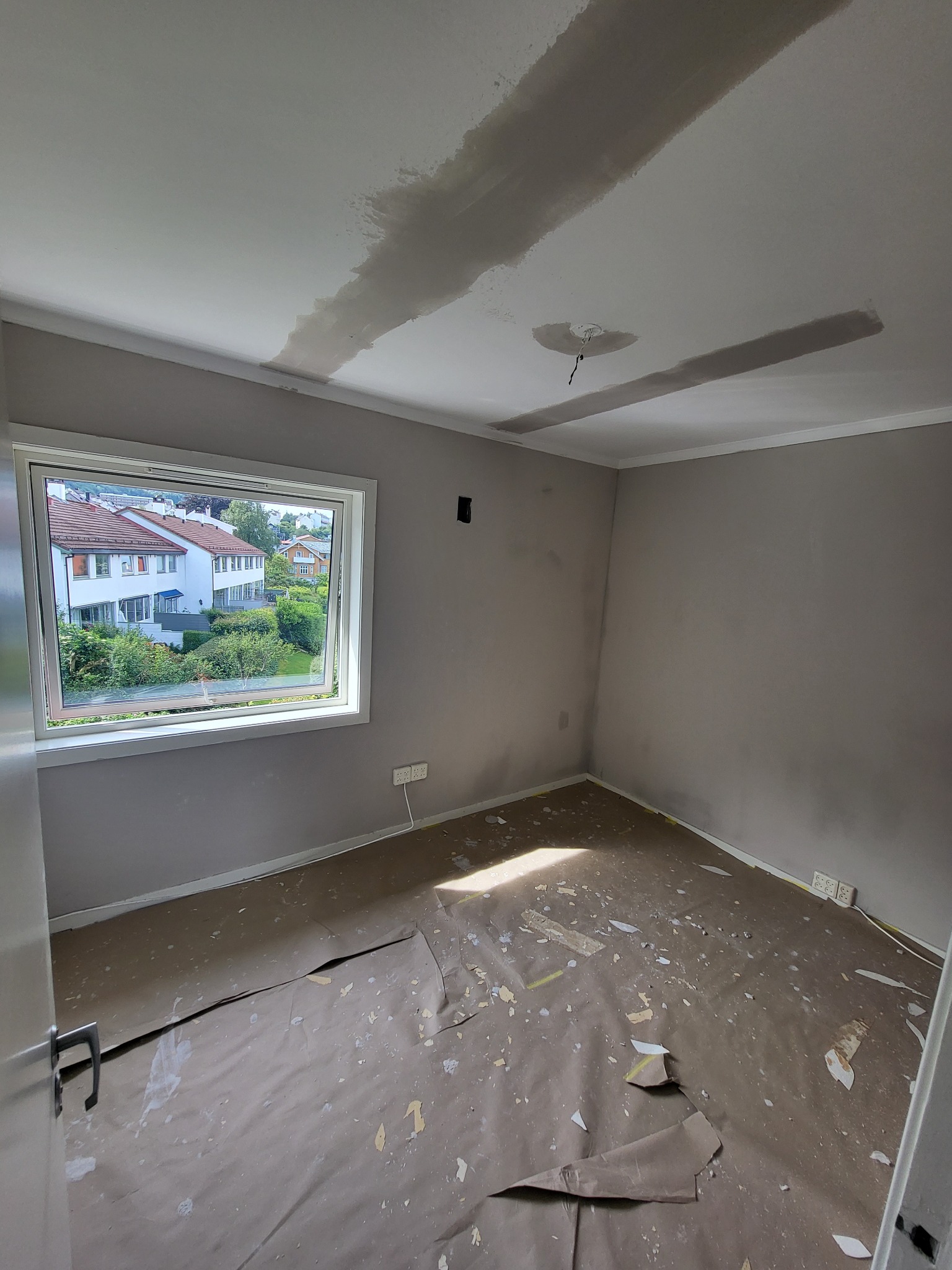 Pokój w trakcie przygotowania do malowania: ściany w szarym odcieniu, zabezpieczona podłoga, widoczne ślady szpachlowania na suficie i wokół otworu na lampę.