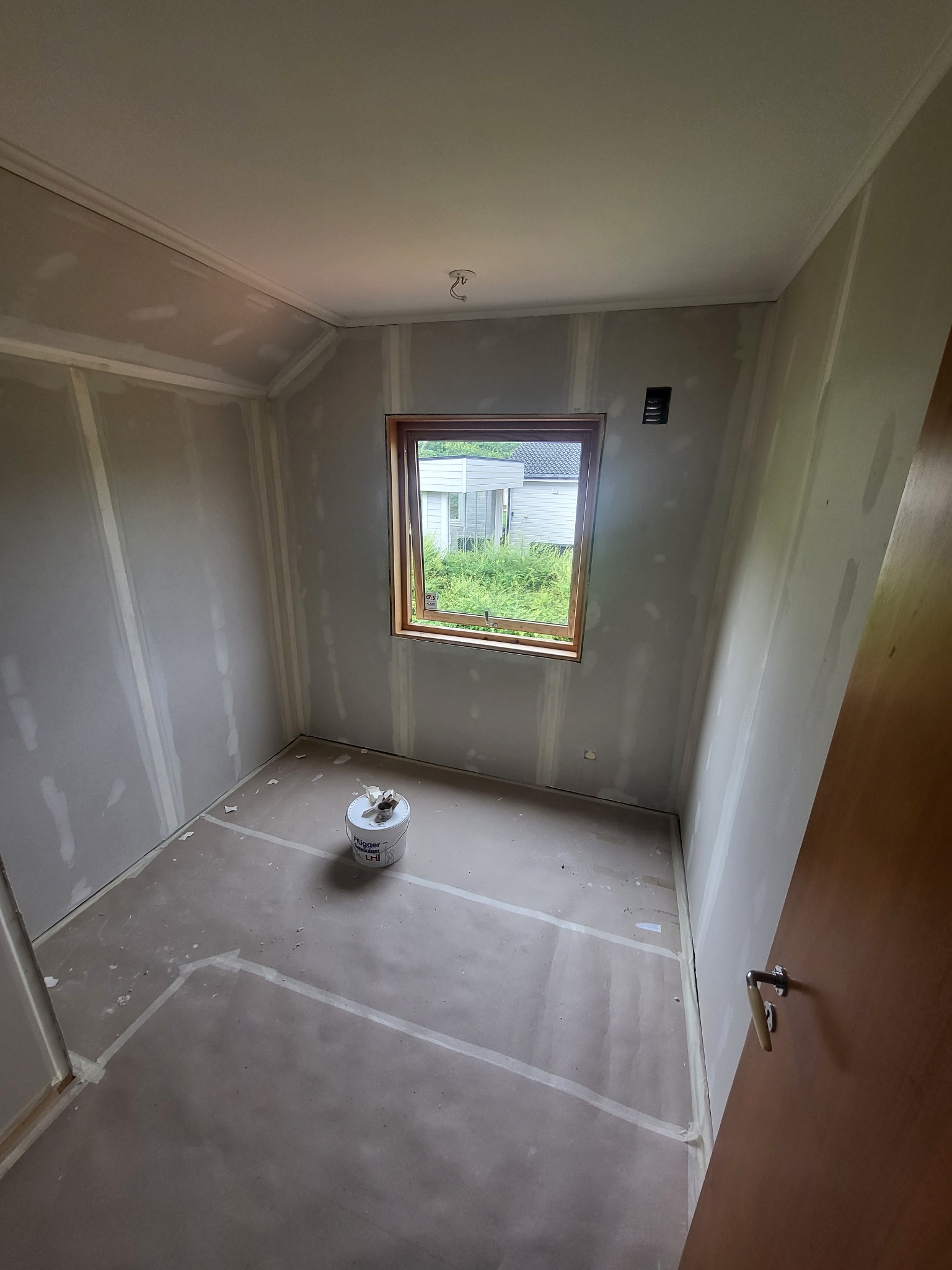 Pokój w trakcie prac wykończeniowych, ściany wyłożone płytami gipsowo-kartonowymi, szpachlowane łączenia, wiadro z farbą na podłodze, widok przez okno na zieleń.