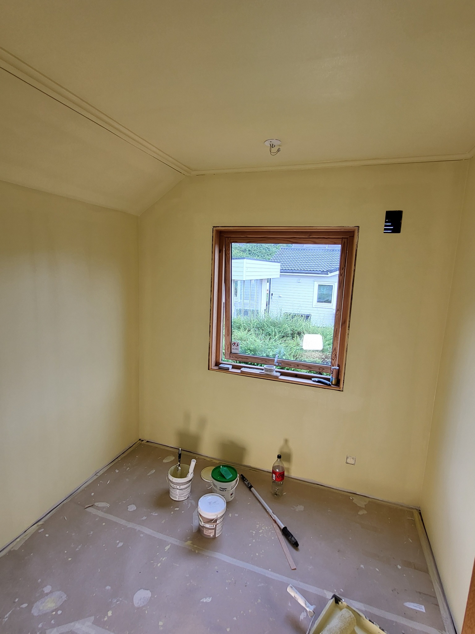 Pokój po malowaniu na żółto, widoczne puszki z farbą, wałek i zabezpieczona podłoga, okno z widokiem na dom z zielenią