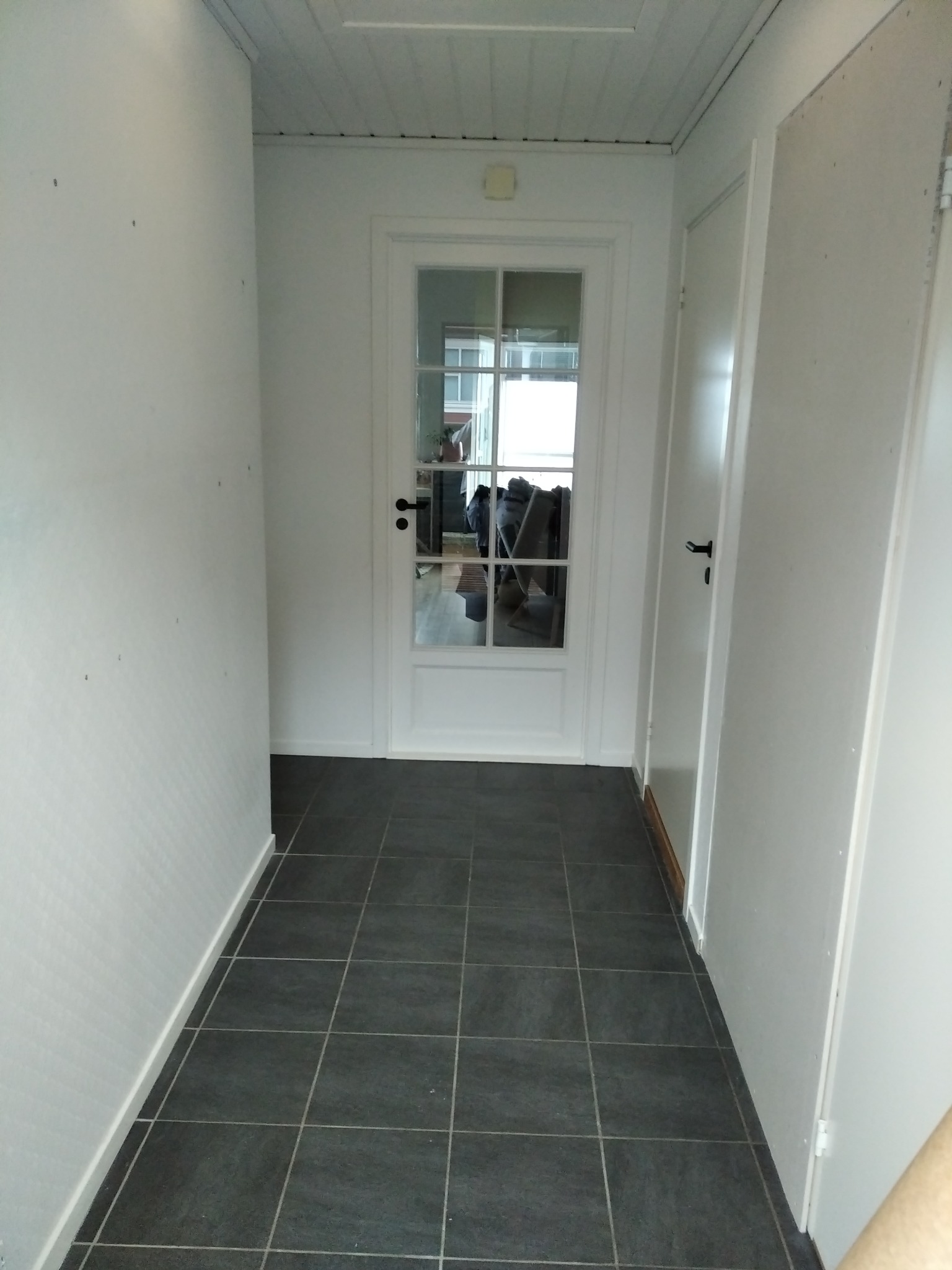 Korytarz z ciemnymi płytkami podłogowymi, białymi ścianami i sufitem z drewnianą boazerią, widok na białe drzwi z szybami i kolejne drzwi po prawej stronie.