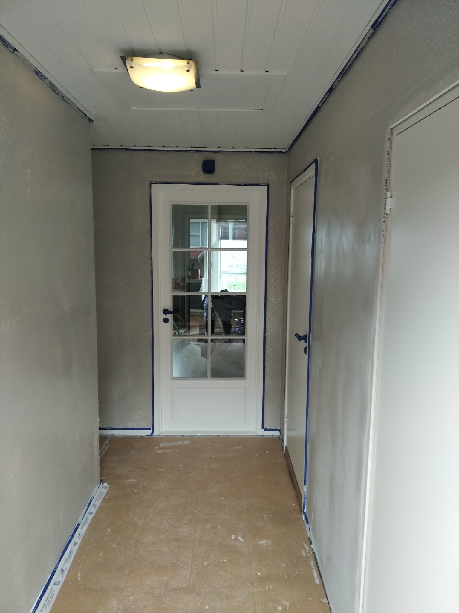 Korytarz w trakcie malowania, ściany zabezpieczone taśmą malarską, widoczna biała stolarka drzwiowa i sufit z panelami.