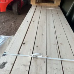 Stos jasnych, heblowanych desek sosnowych, spiętych białą taśmą, na palecie, z widocznym w tle wózkiem widłowym.