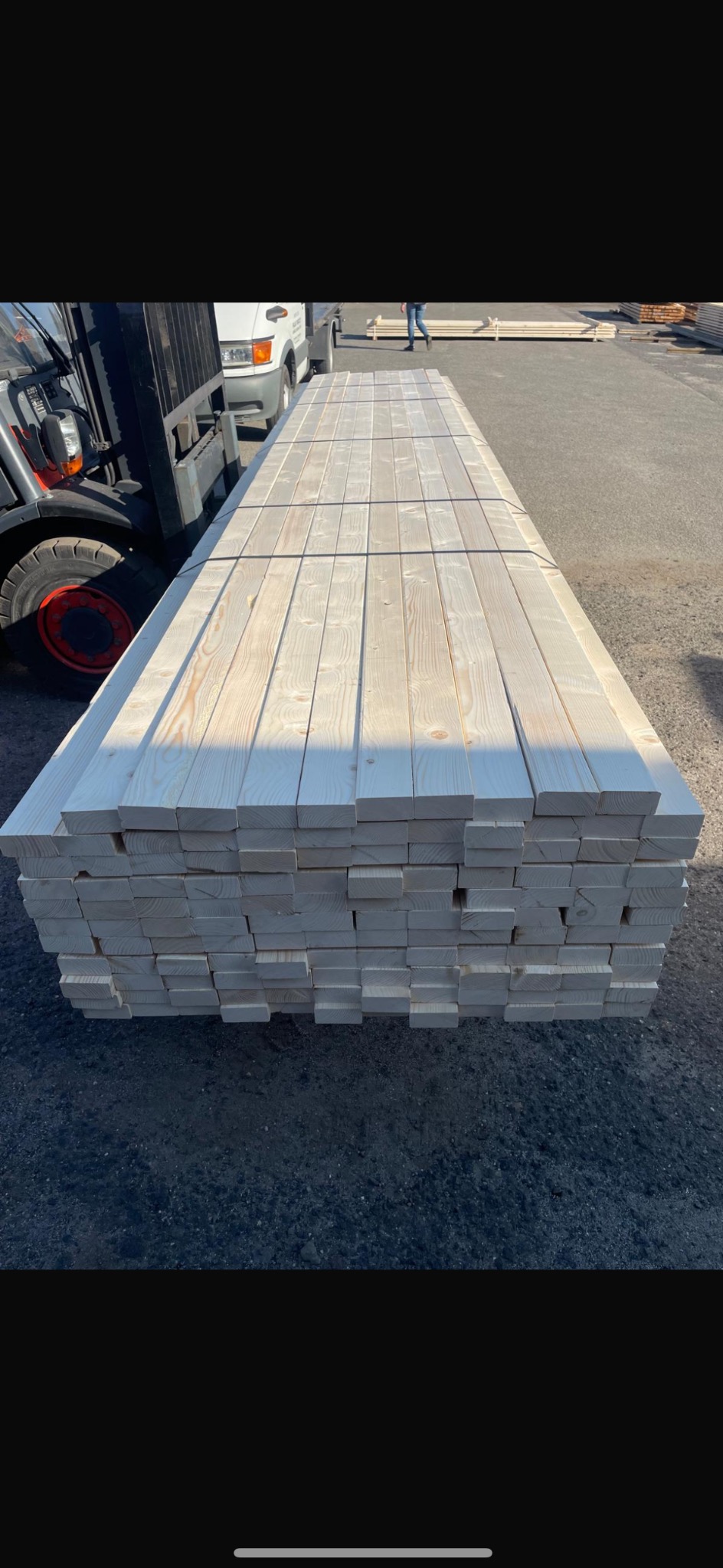 Stos jasnej, świeżo obrobionej tarcicy budowlanej, zabezpieczony taśmami, leżący na placu, z wózkiem widłowym i samochodem dostawczym w tle.