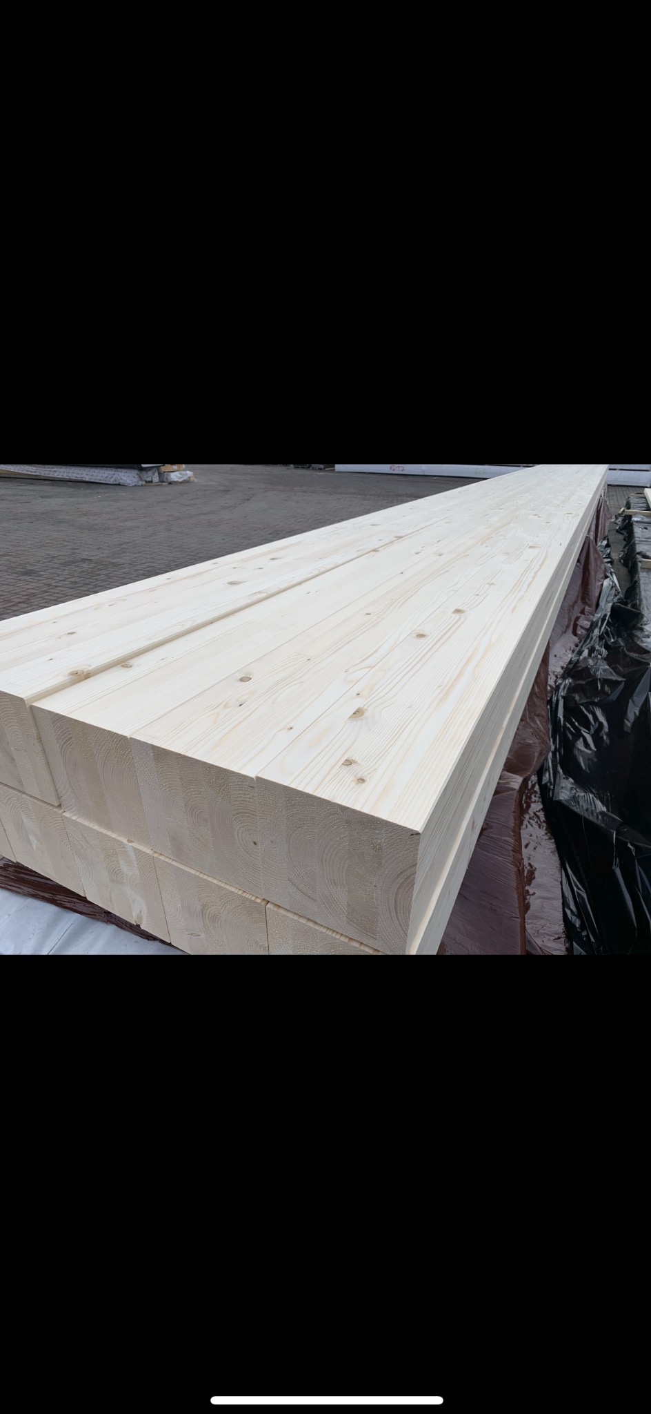 Stos jasnych, heblowanych belek drewnianych, widoczny przekrój z wyraźnym rysunkiem słojów, zabezpieczonych folią na zewnątrz.