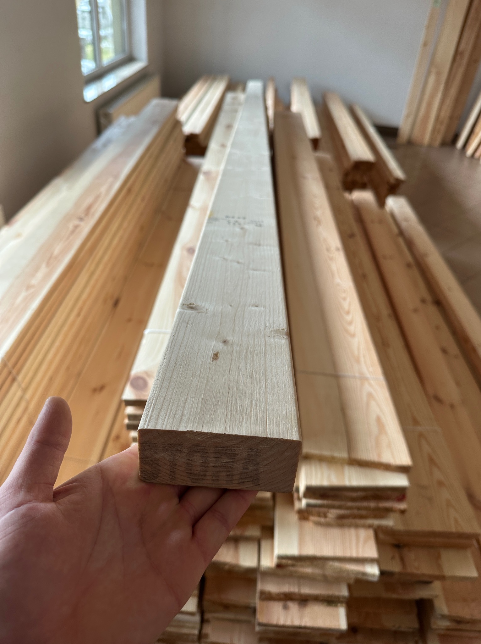 Ręka trzymająca kantówkę z jasnego drewna na tle stosu tarcicy budowlanej w pomieszczeniu.