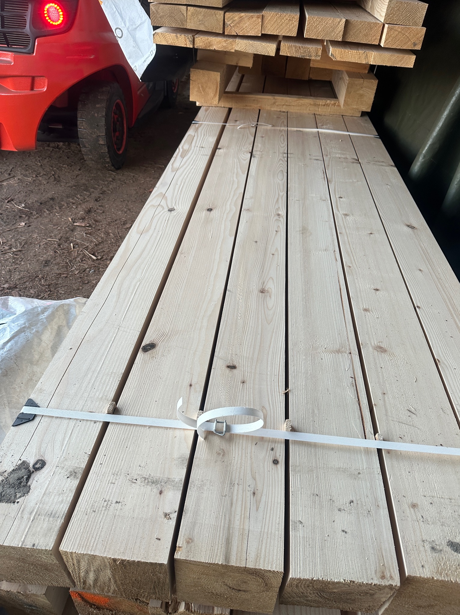 Stos jasnych, heblowanych desek sosnowych, spiętych białą taśmą, na palecie, z widocznym w tle wózkiem widłowym.