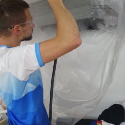 Strefa AC Sławomir Smoter - Mężczyzna w okularach ochronnych czyści klimatyzator zawieszony pod sufitem, używając specjalnego urządzenia z dyszą, całość zabezpieczona folią ochronną.